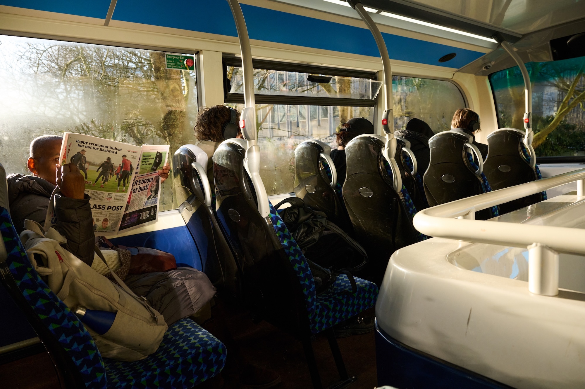 Interior of bus with blue and white patterned seats, passengers seated throughout, bright sunlight streaming through windows.