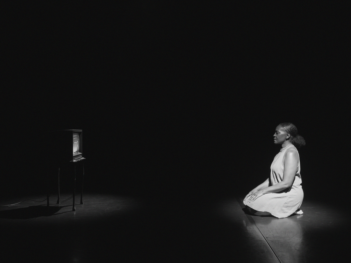 Black and white image: woman in light dress kneeling on floor in dark room, illuminated monitor on left side, dramatic lighting.