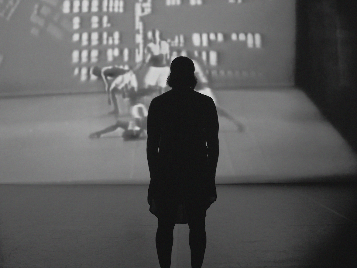 Person silhouetted in dark room viewing projected film or video on wall showing figures and text in black and white.