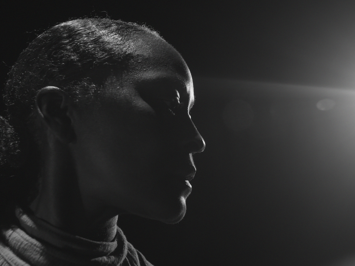 Black and white profile of man with short hair looking right, dramatic side lighting creates strong shadows against dark background.