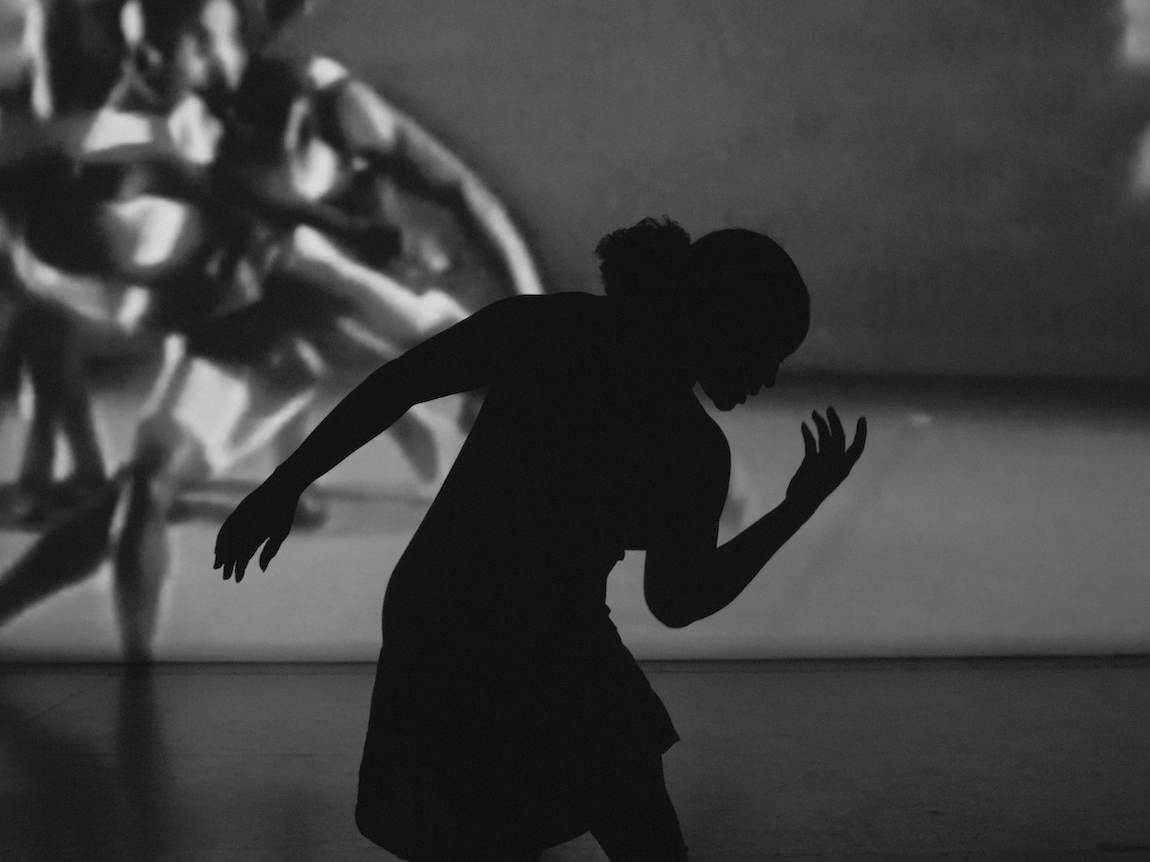 Black silhouette of person with arms extended against grey wall with projected shadows in background. Monochrome image.