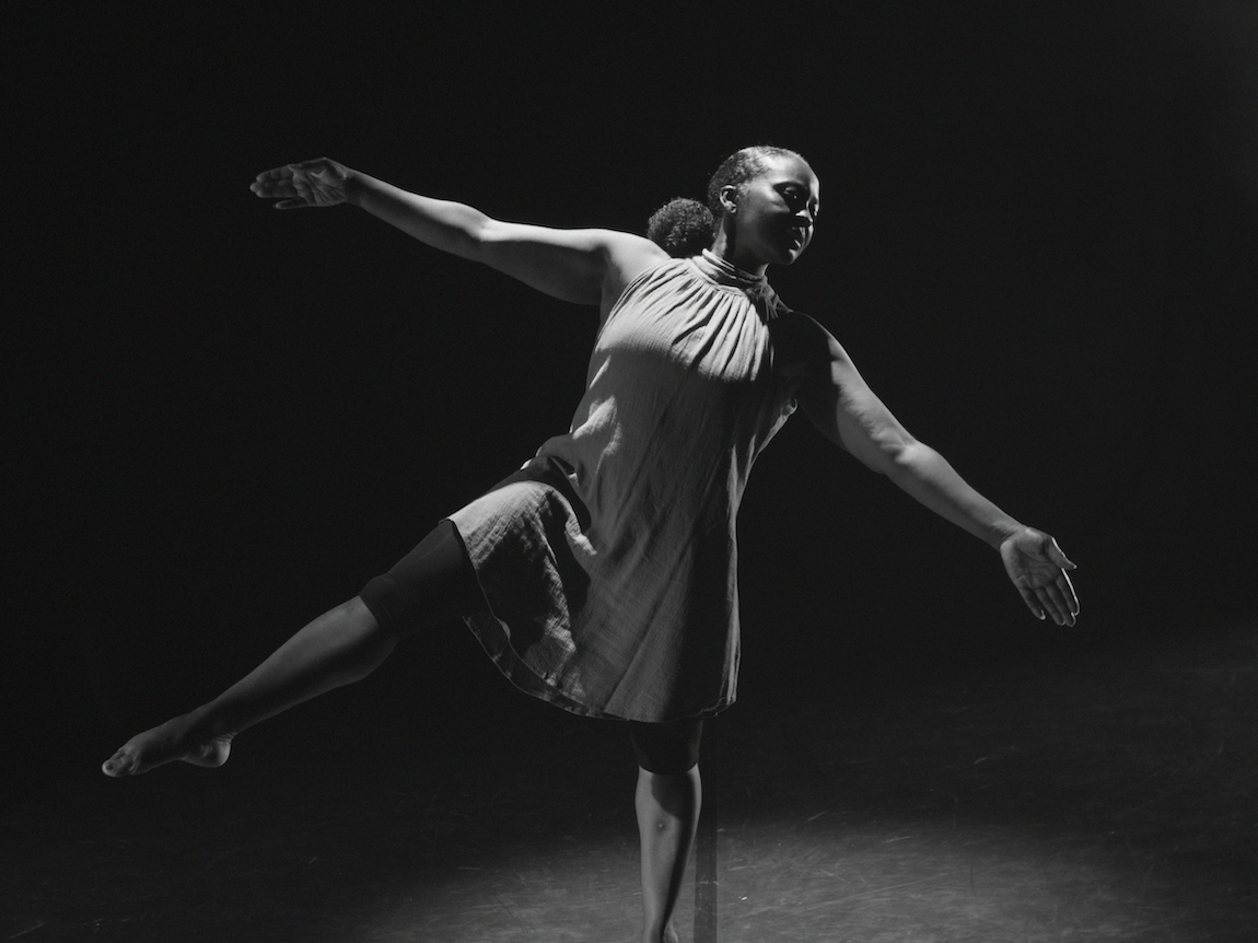 Black and white image of woman in flowing dress with arms outstretched and one leg extended, captured mid-dance movement against dark background.