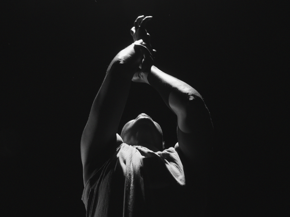 Black and white image of dancer in flowing fabric against dark background, arms raised above head in graceful pose.
