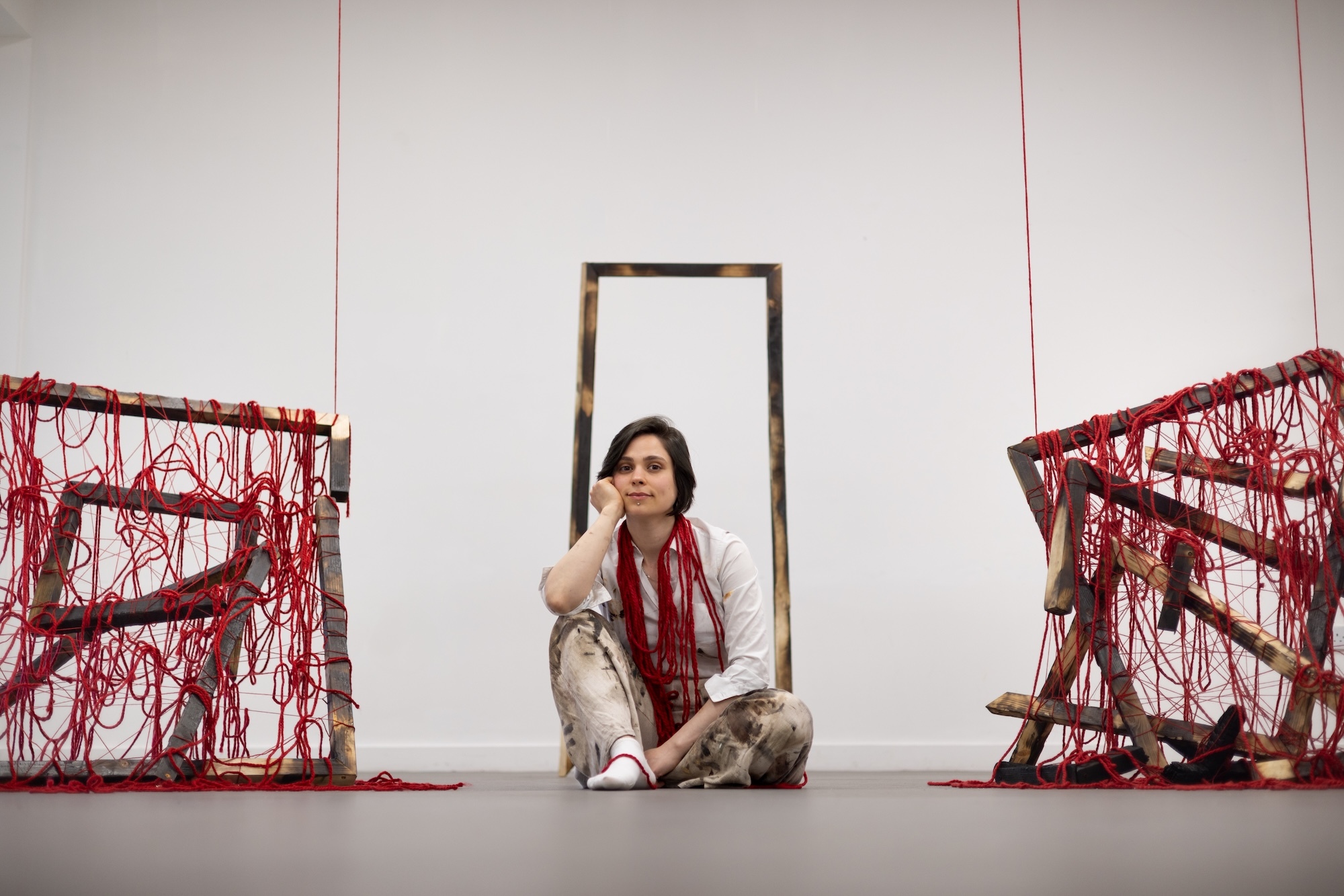S Adi sitting between two red string installations, with framed artwork on white wall behind.