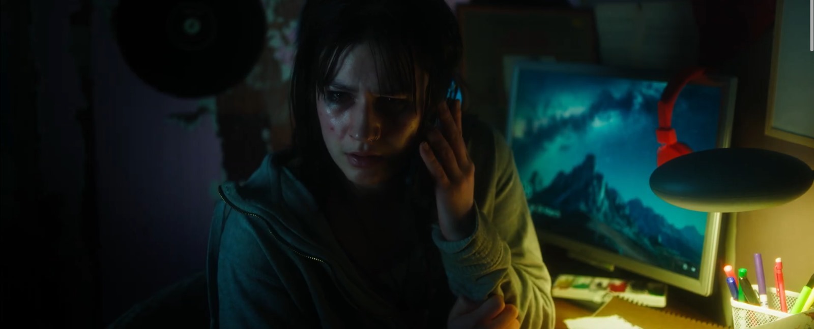 Person with long dark hair in yellow jacket sitting at desk with computer monitor displaying blue screen in dimly lit room.