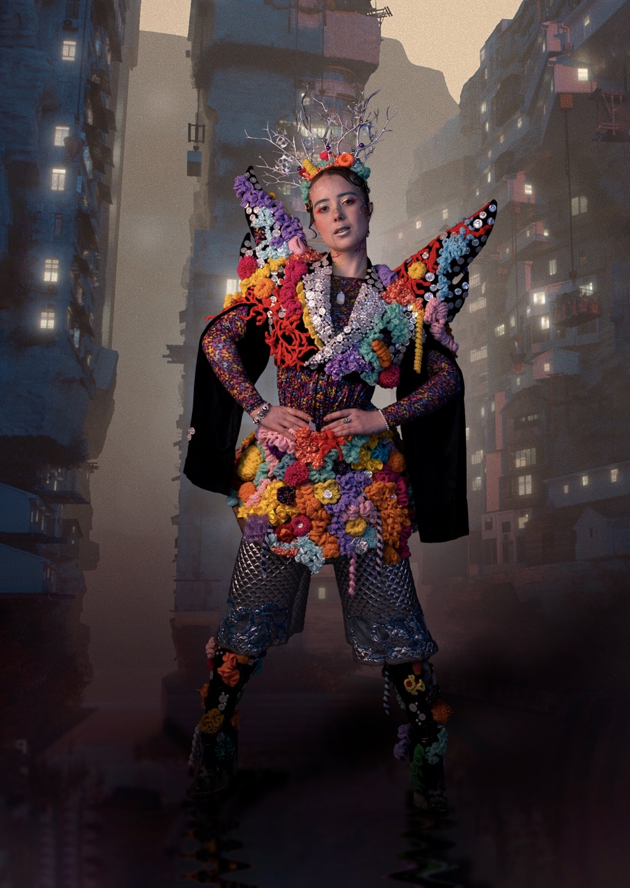 A person wearing an elaborate, brightly coloured floral costume stands in an urban alley with tall buildings in the background.