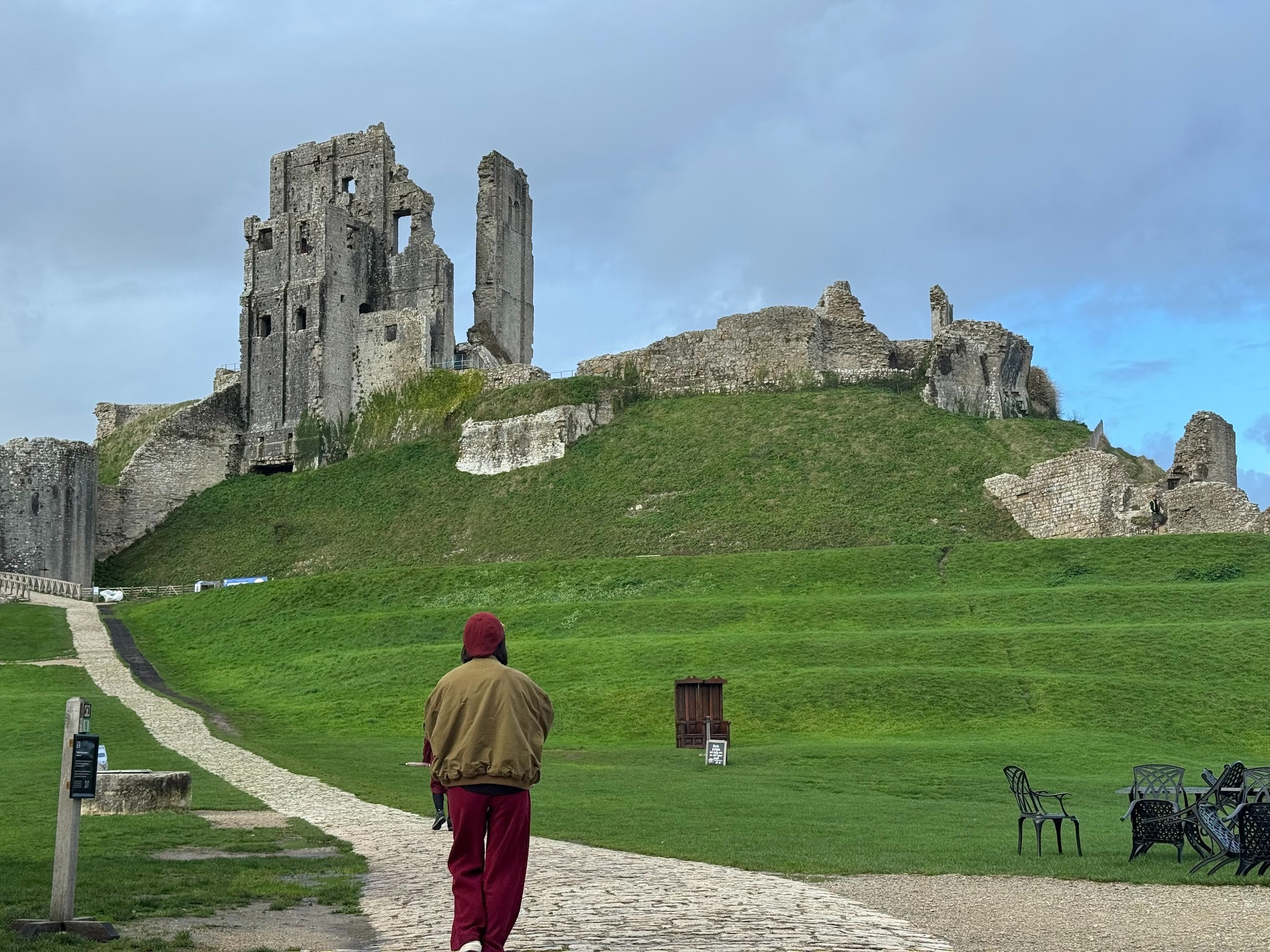 A person in a red hooded sweatshirt walks along a stone pathway towards the ruins of a castle on a grassy hill. The landscape features uneven terrain, a few benches, and remnants of stone walls under a partly cloudy sky.