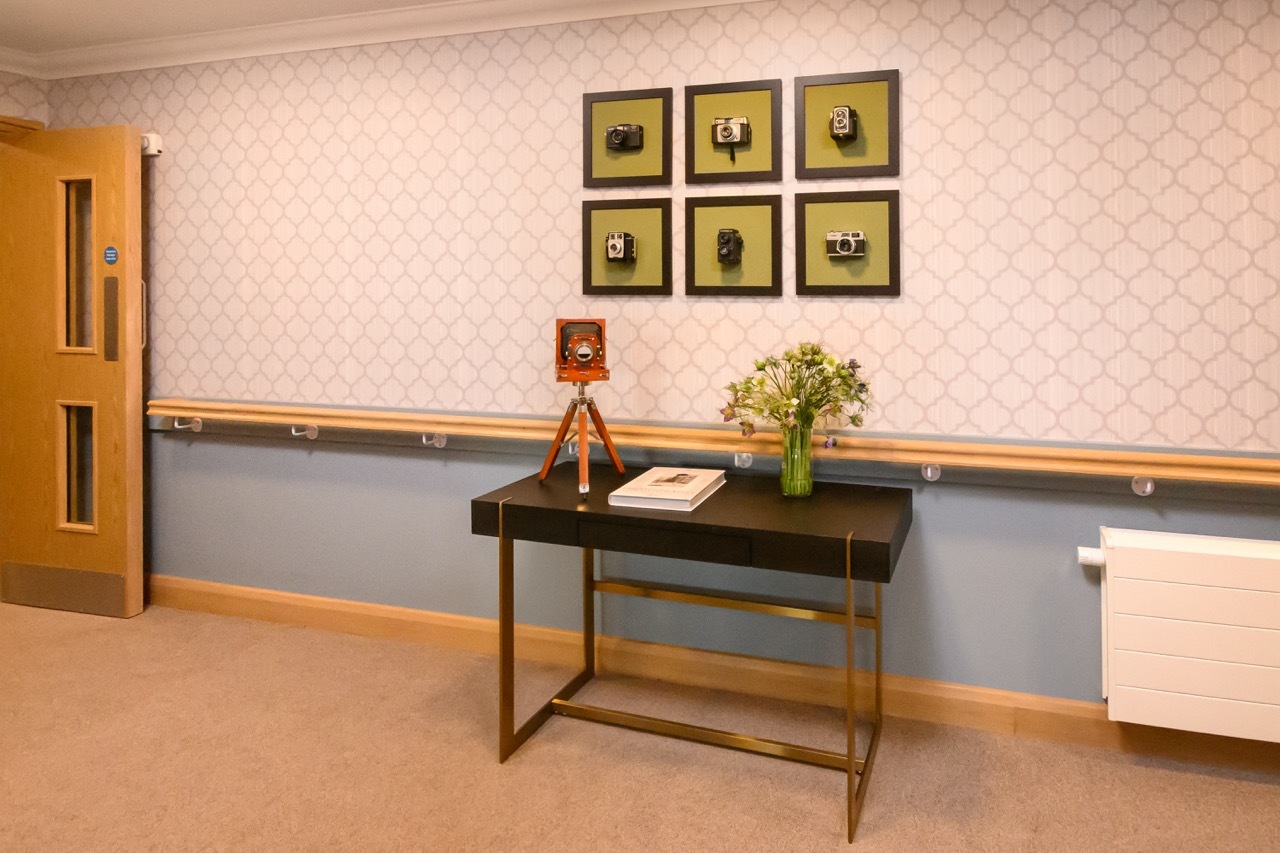 A corridor in Diana Celella's 'Wellford Gardens Care Home' with a stylised table and old cameras on display and framed.
