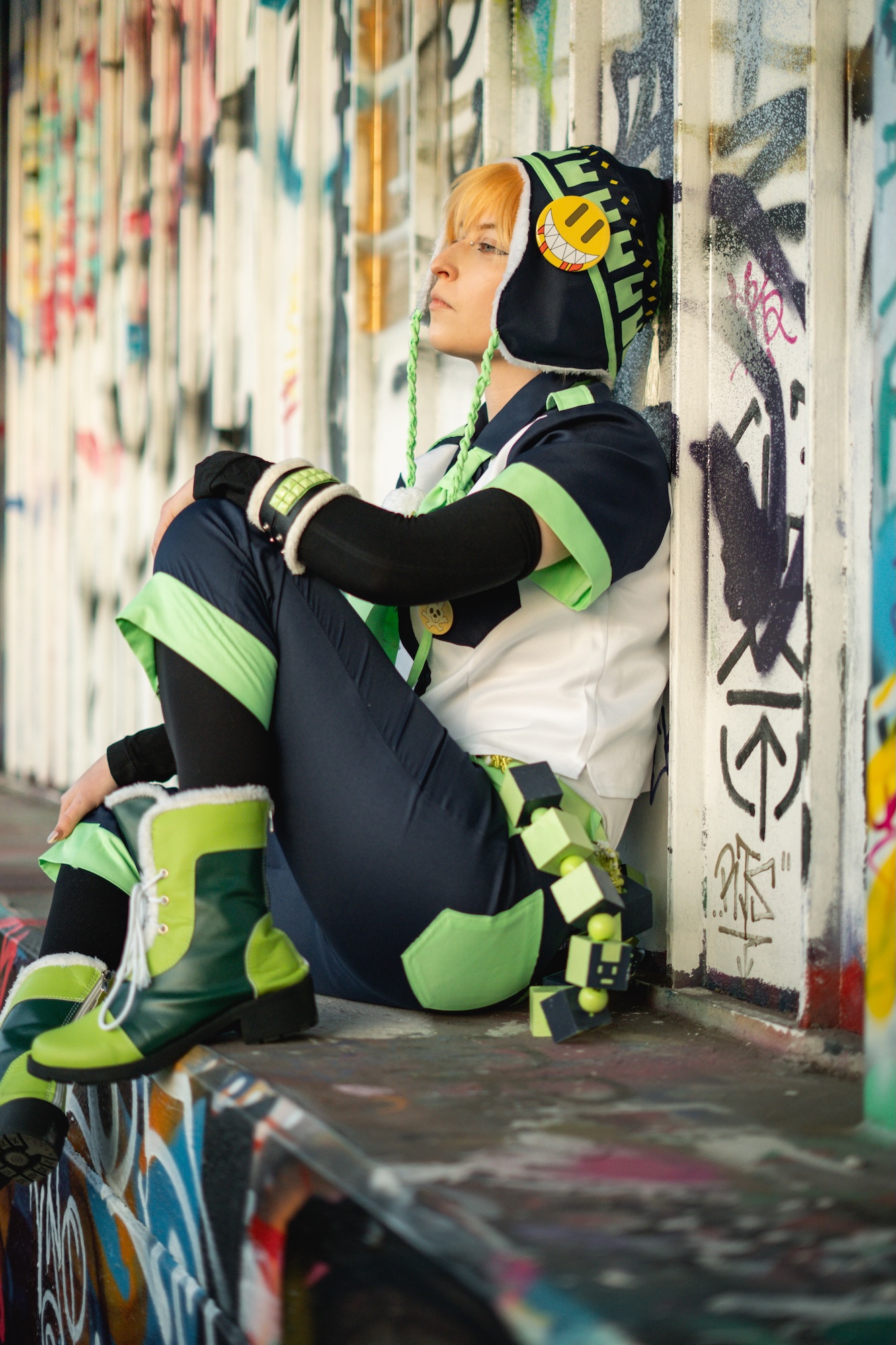 A person sits against a graffiti-covered wall, wearing a green and black outfit, with a black beanie and headphones. Their legs are crossed, and they are looking to the side. The background features vibrant street art in various colours.