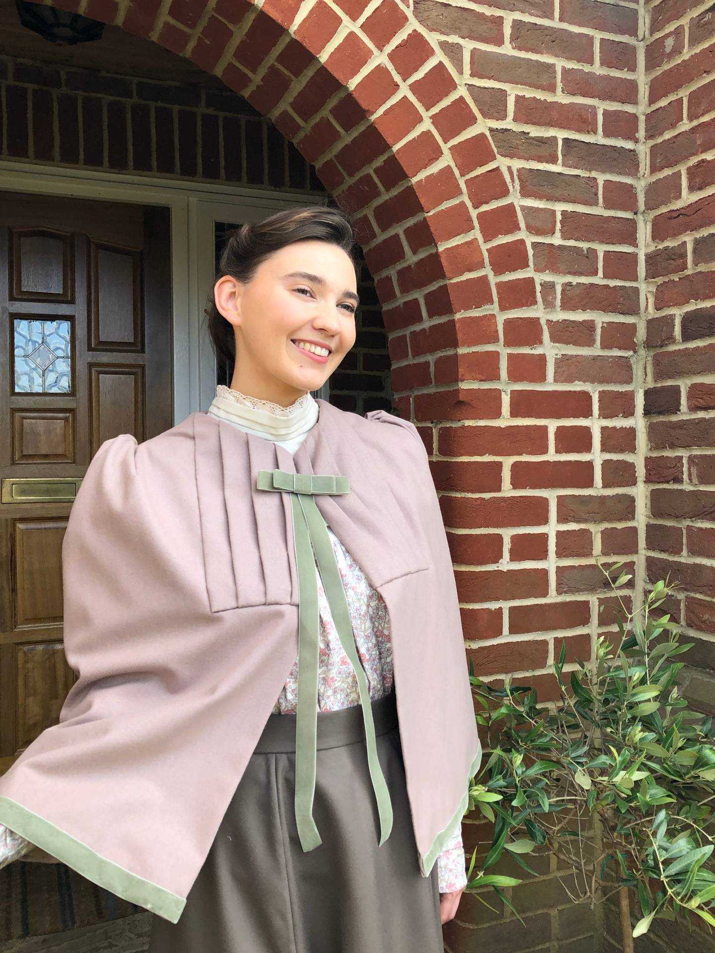 A person in a period costume stands smiling outside a house with a brick archway at the front.