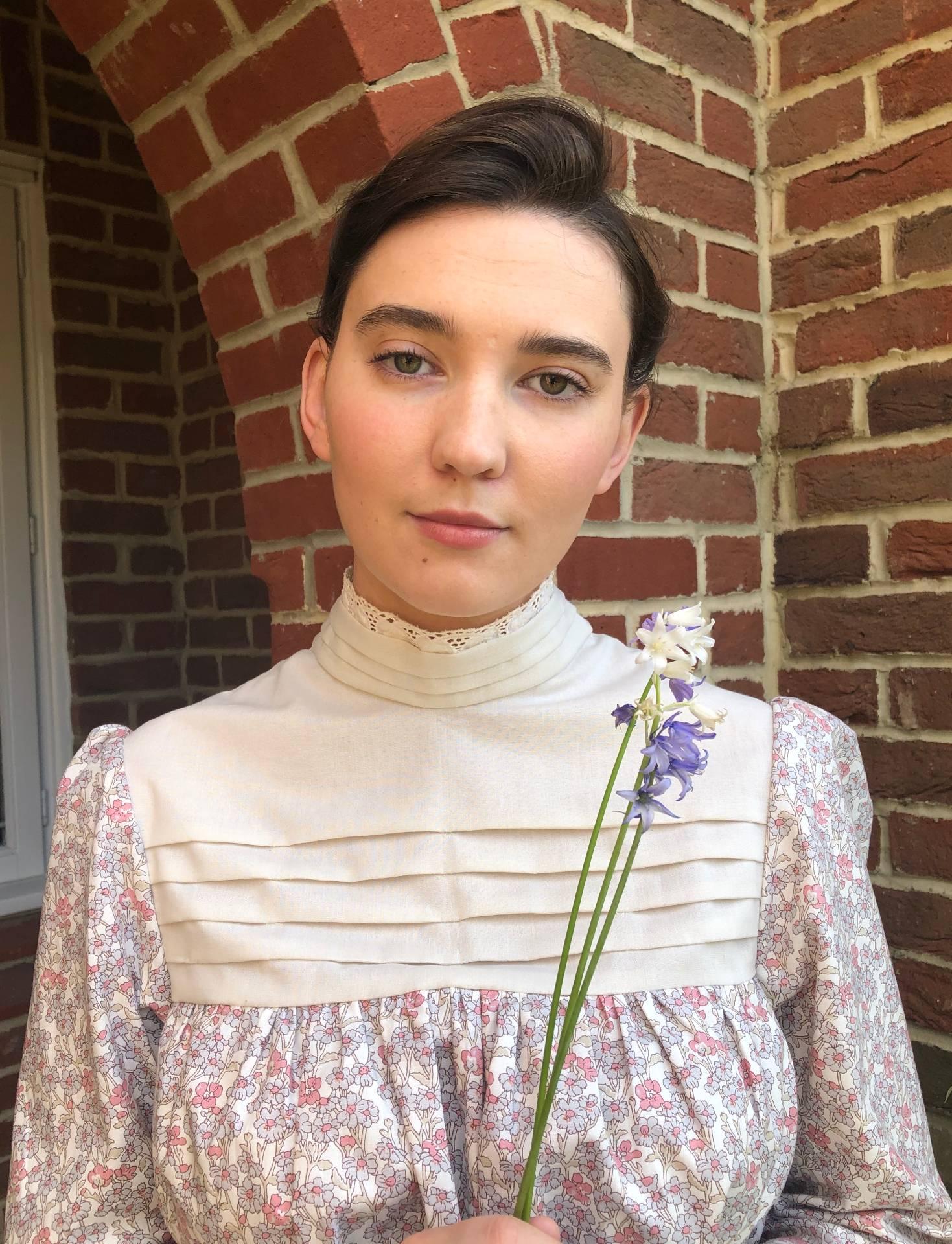 A person in period costume stands outside a house holding some flowers.