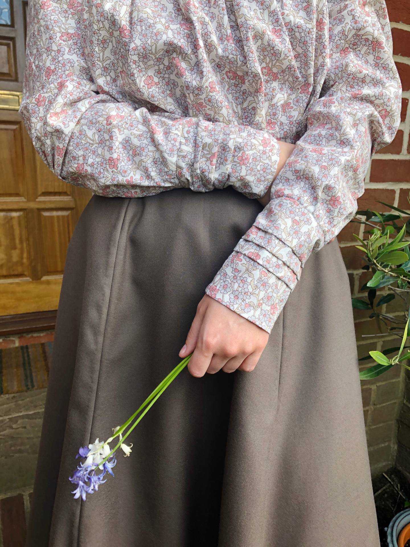 The torso of a person wearing a period costume and their arms crossed, holding bluebells in one hand.