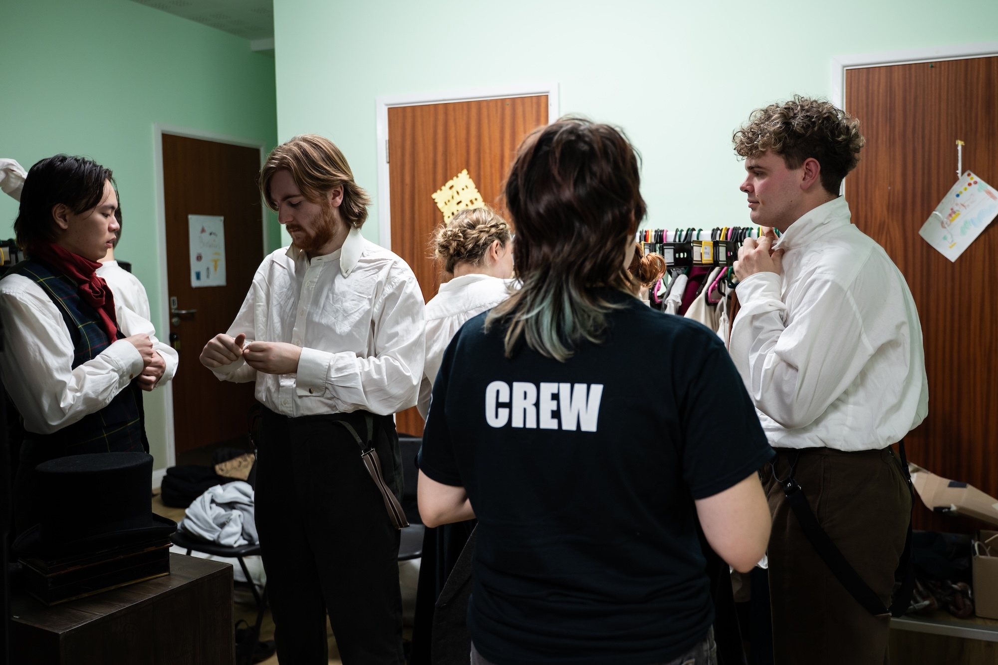 BA (Hons) Acting and BA (Hons) Costume students in white shirts and one person in black "CREW" t-shirt standing in green-walled room with wooden cabinets and bottles.