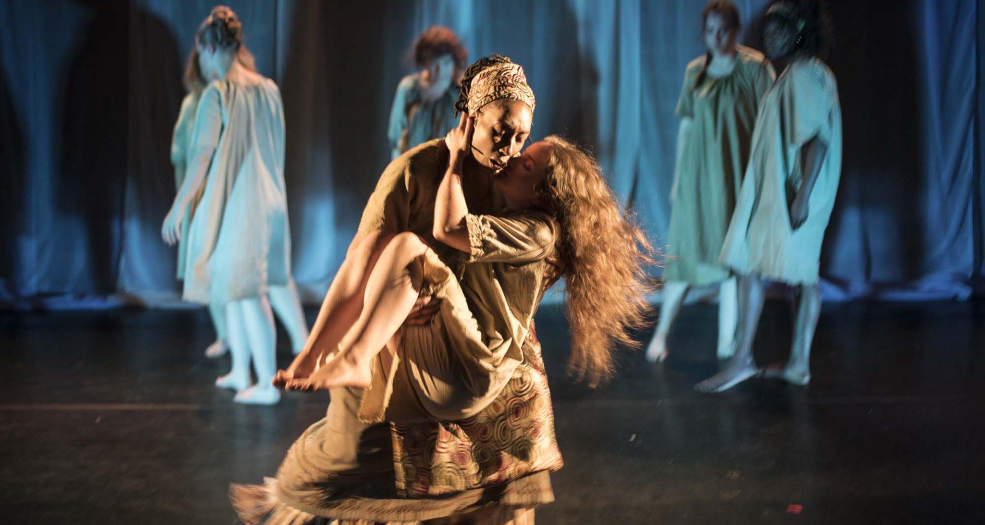 Two acting students on stage in costume in the middle of a performance, holding each other. A small group of other performers appear in the background.