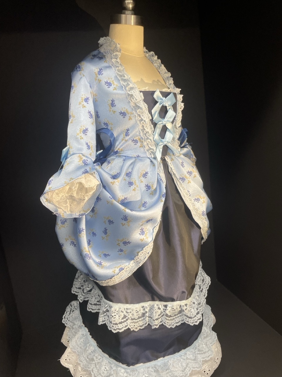 Blue floral dress with white lace trim and blue bows, dark background.