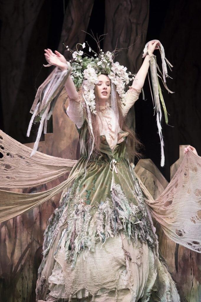 A person in a ragged fairytale-style costume and floral headdress raises her arms up.