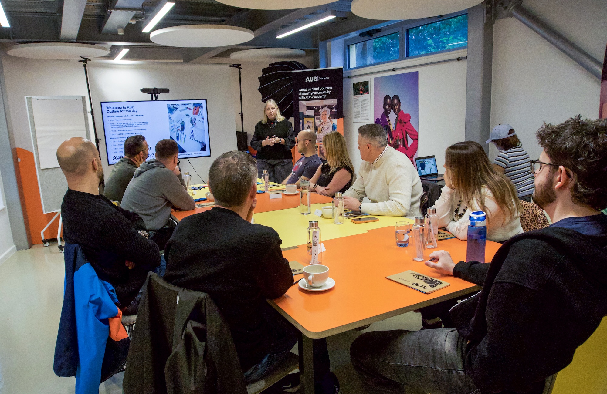 A group of people seated around an orange conference table, watching a woman present beside a screen in a meeting room with posters and large windows.