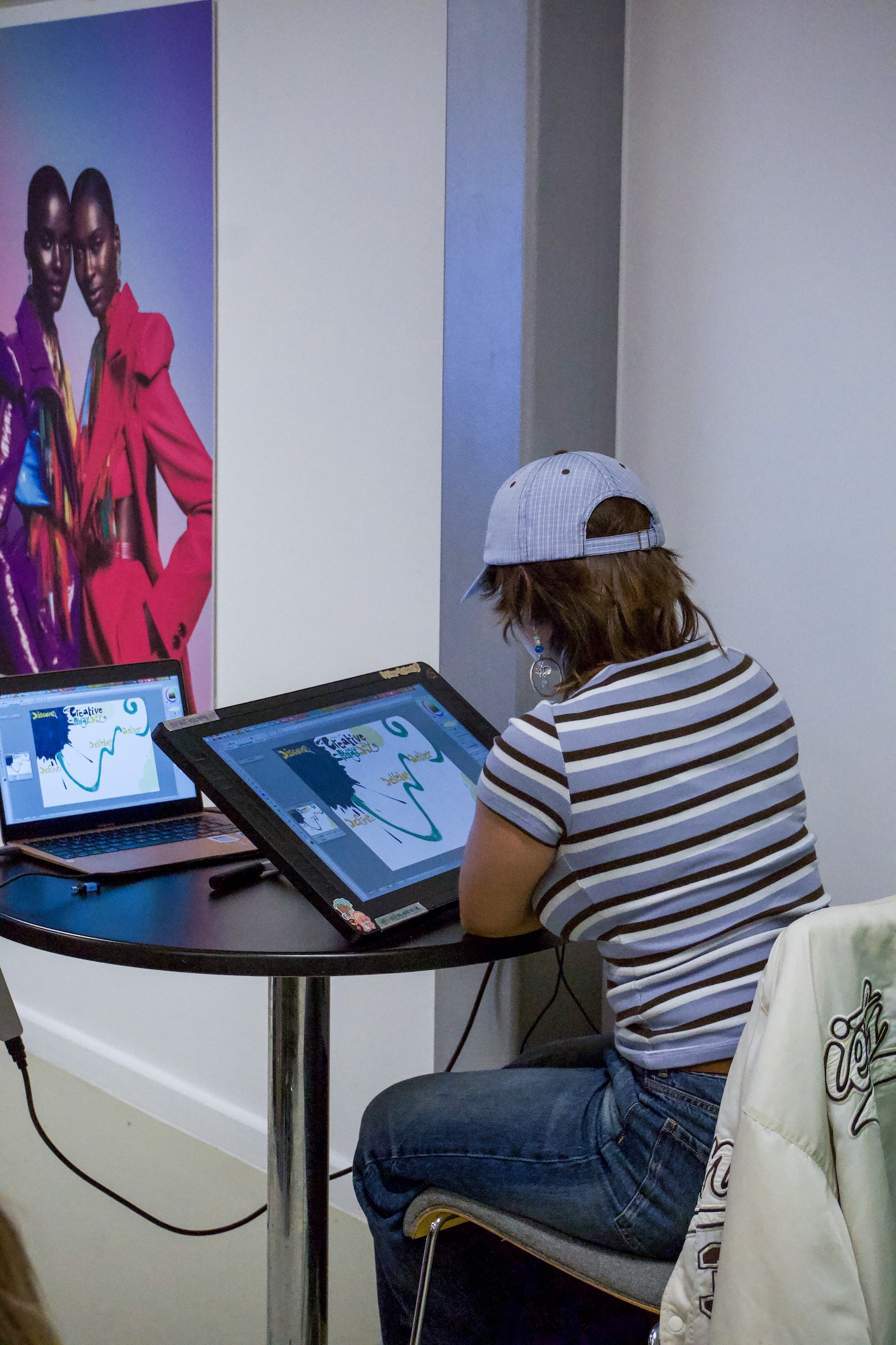 A person in a white cap and striped T-shirt uses a digital drawing tablet at a round black table, with a laptop, wall poster and plain white walls in the background.