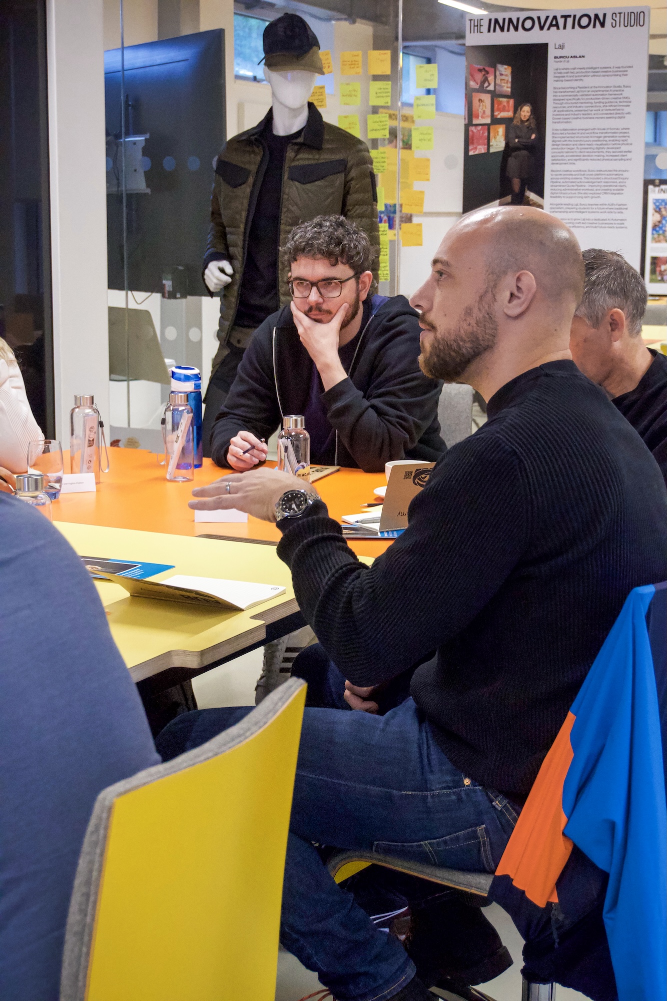 Three men sit around a table in a workshop area, with a mannequin and an ‘Innovation Studio’ display in the background.