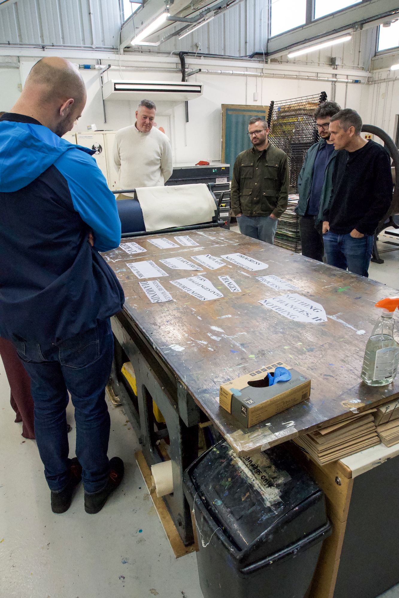 Four men stand around a large worktable covered with paper labels in a workshop with metal shelving and tools.