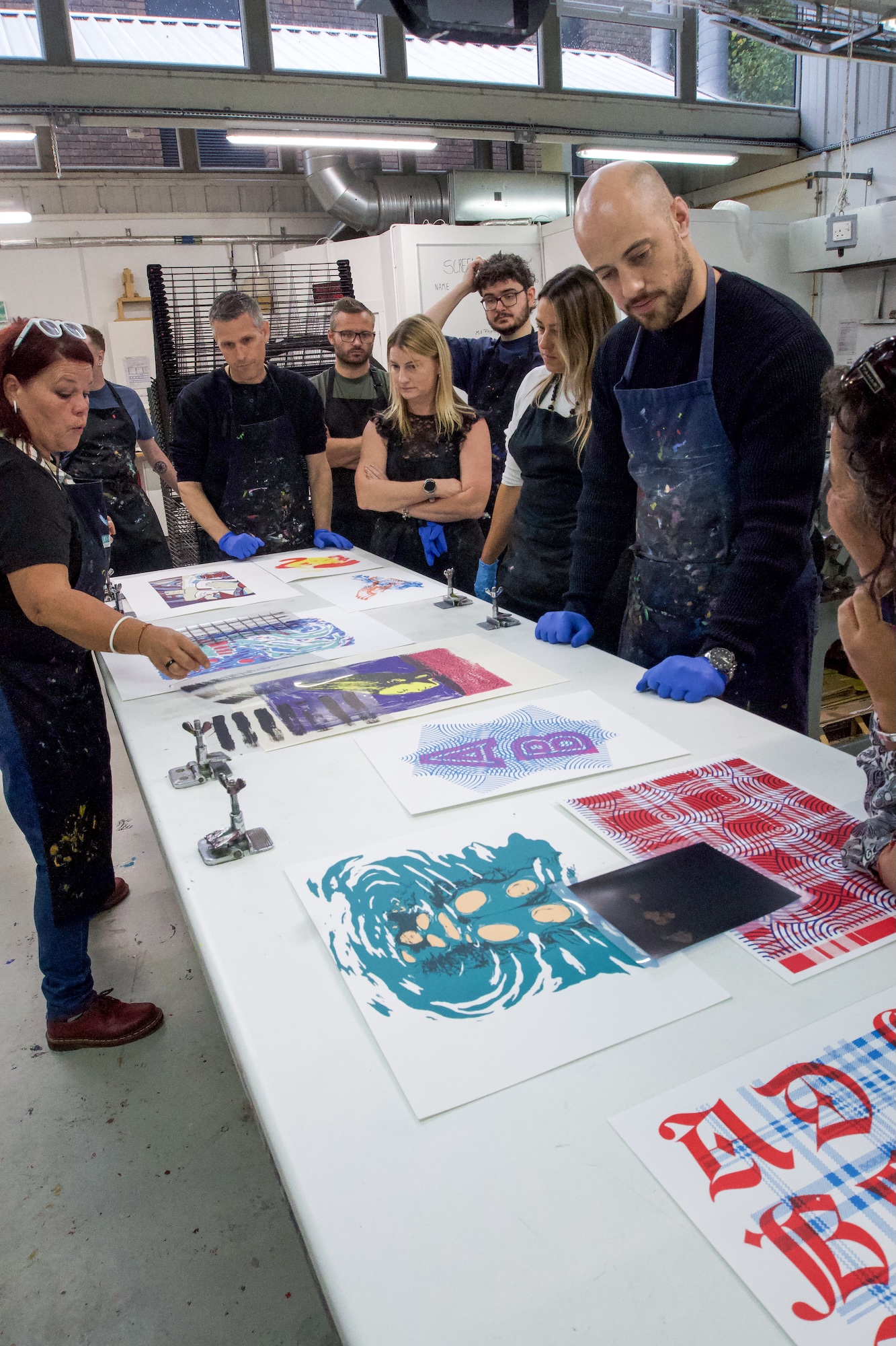 A group of people wearing gloves stands around a large table in a print workshop, looking at colourful printed designs and posters laid out on the surface.