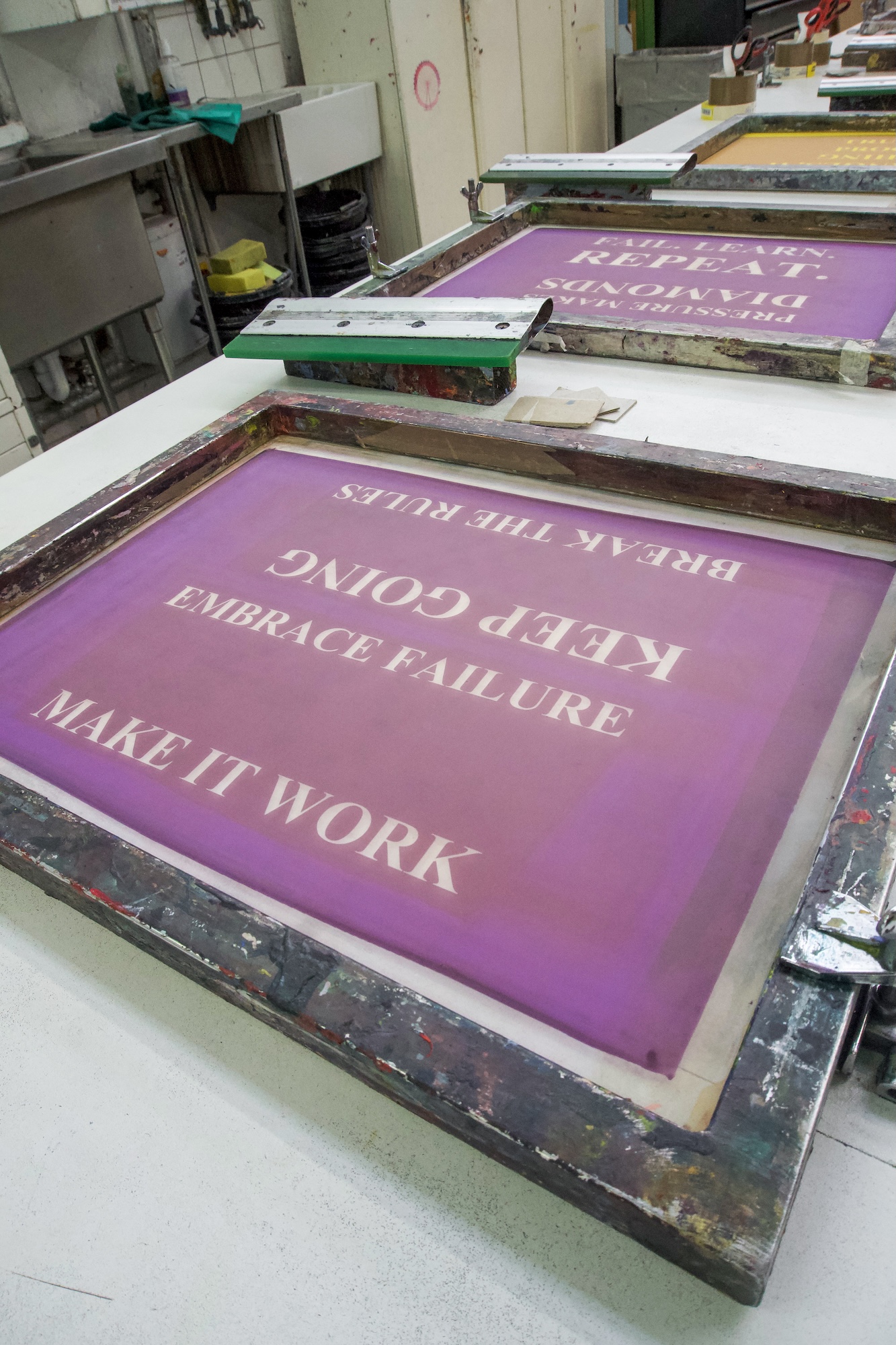 A screen-printing frame with a purple poster reading “KEEP GOING, EMBRACE FAILURE, MAKE IT WORK” on a workshop table.
