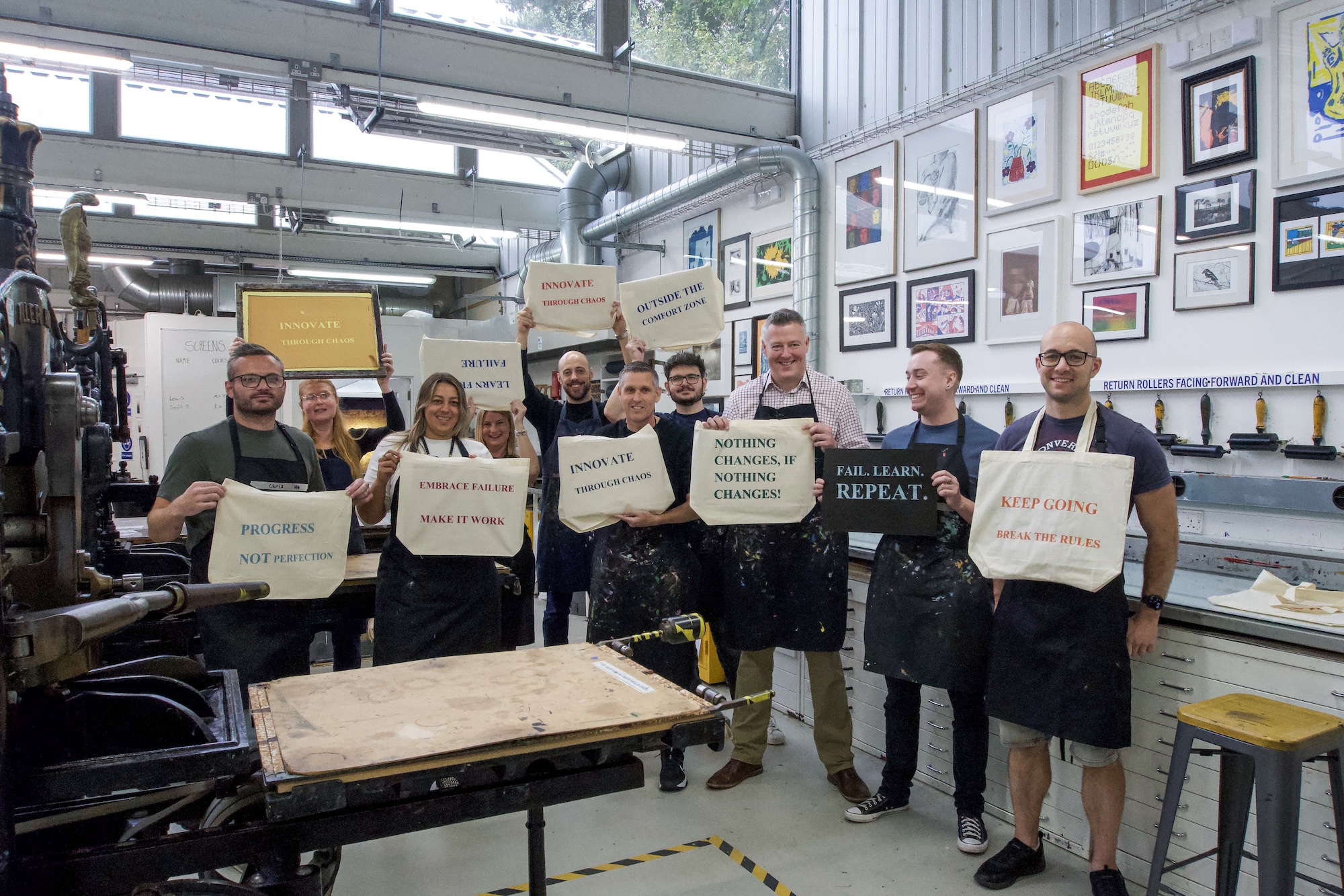 Eight people standing in a print workshop, holding placards with motivational slogans beside a large printing press and framed artwork on the wall.