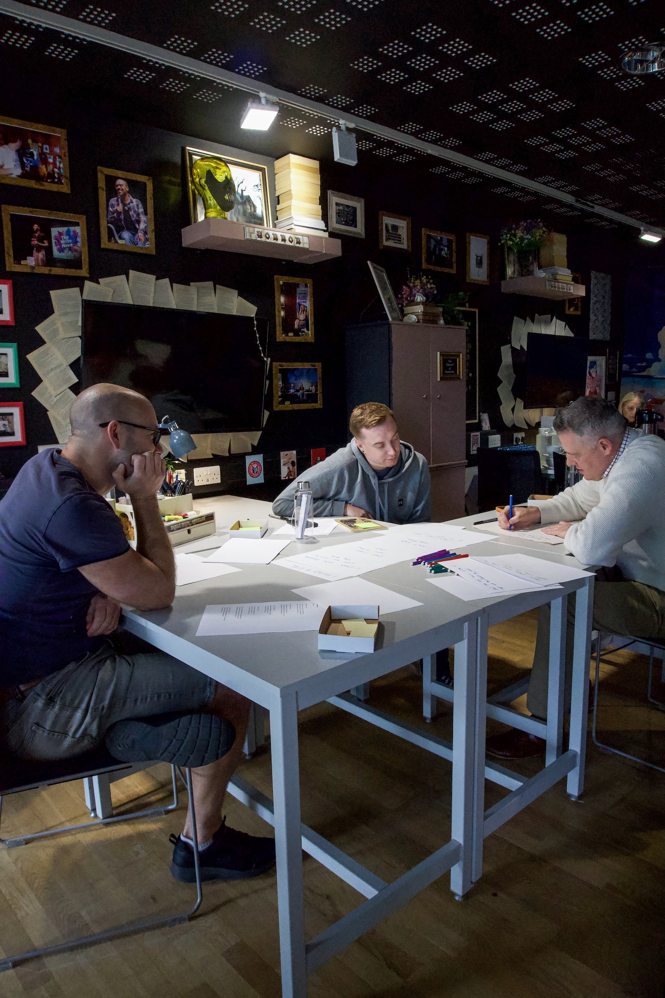 Three men sit around a white table covered with papers and markers in a dark room, with framed photographs and sketches displayed on the walls.