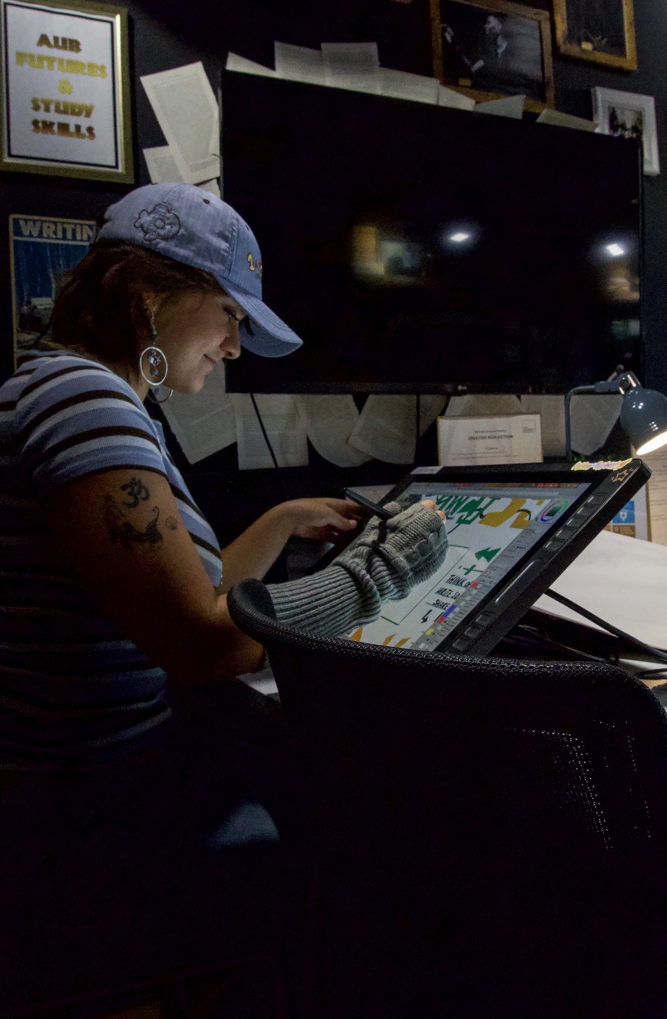 A woman wearing a blue cap and striped shirt works on a digital drawing tablet beside a large monitor in a dim studio, with framed posters on the wall.