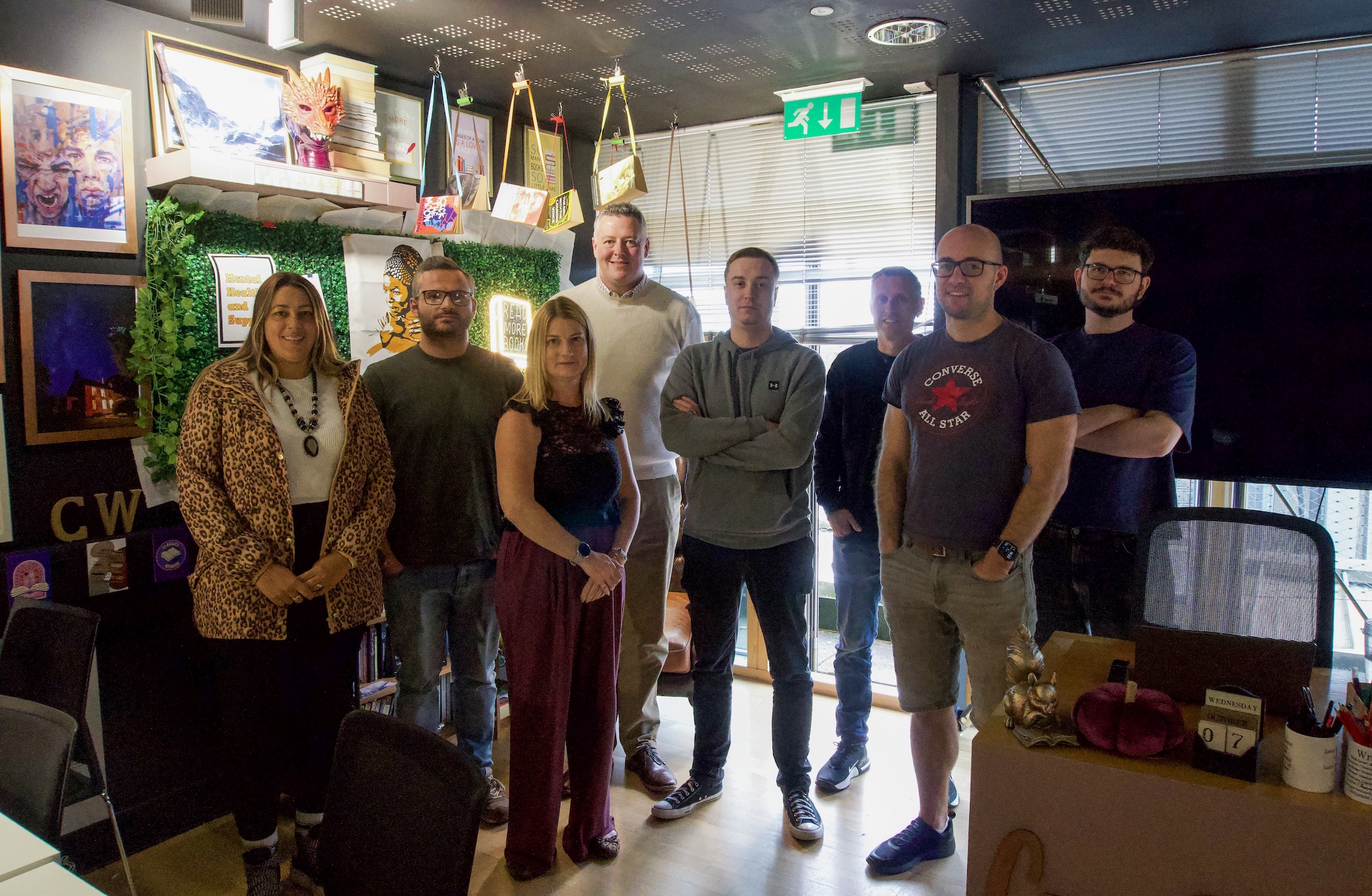Seven people stand together in a small office, with posters, plants and computer equipment around them.