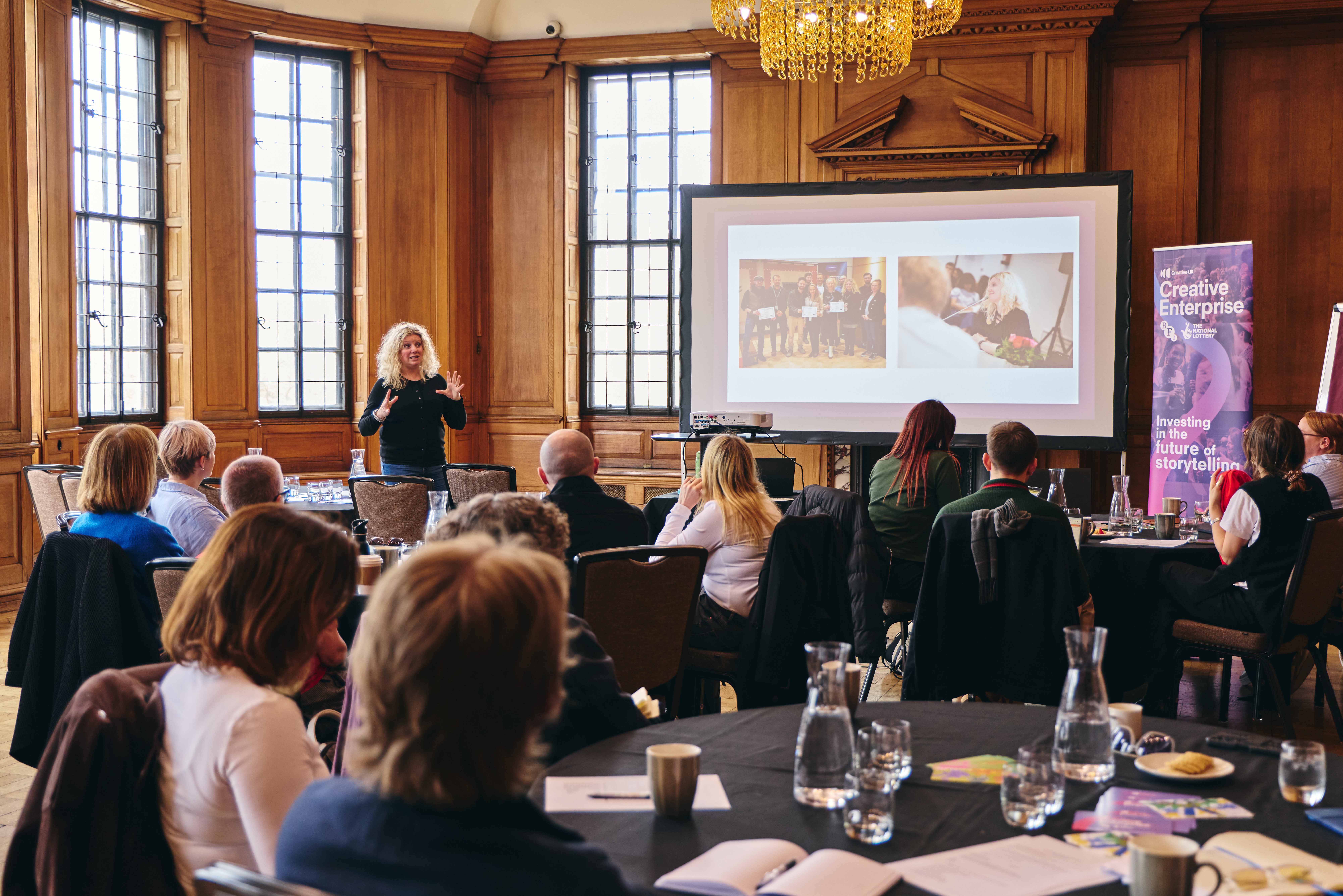 People gathered in a wooden wall space listening to a Creative Enterprise talk, slides projected and woman with blonde hair talking.