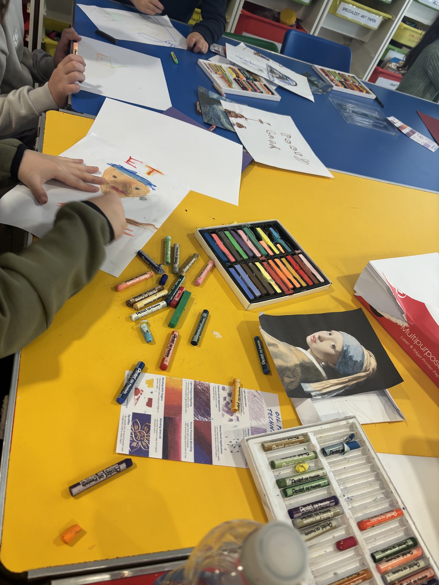 A colourful table covered with various art supplies, including oil pastels, papers, and a water bottle. Children’s hands are seen drawing on white sheets, while a book with a famous painting is placed next to the art materials.