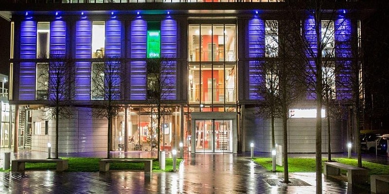 Front view of a well-lit university building with a wet pavement and lawn outside.