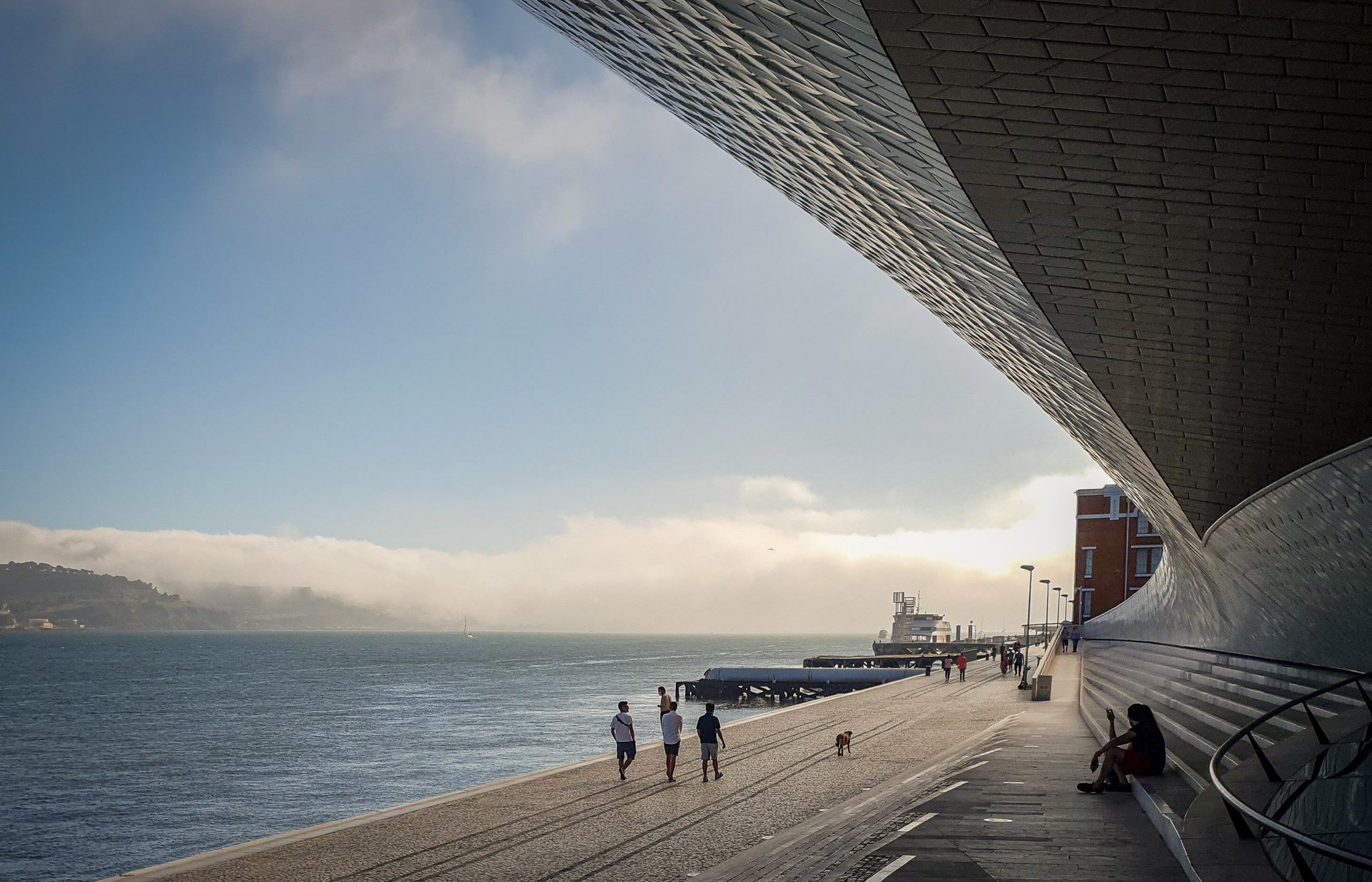 A promenade by the sea. Three people and a dog walk along. Someone sits on some steps to the right.