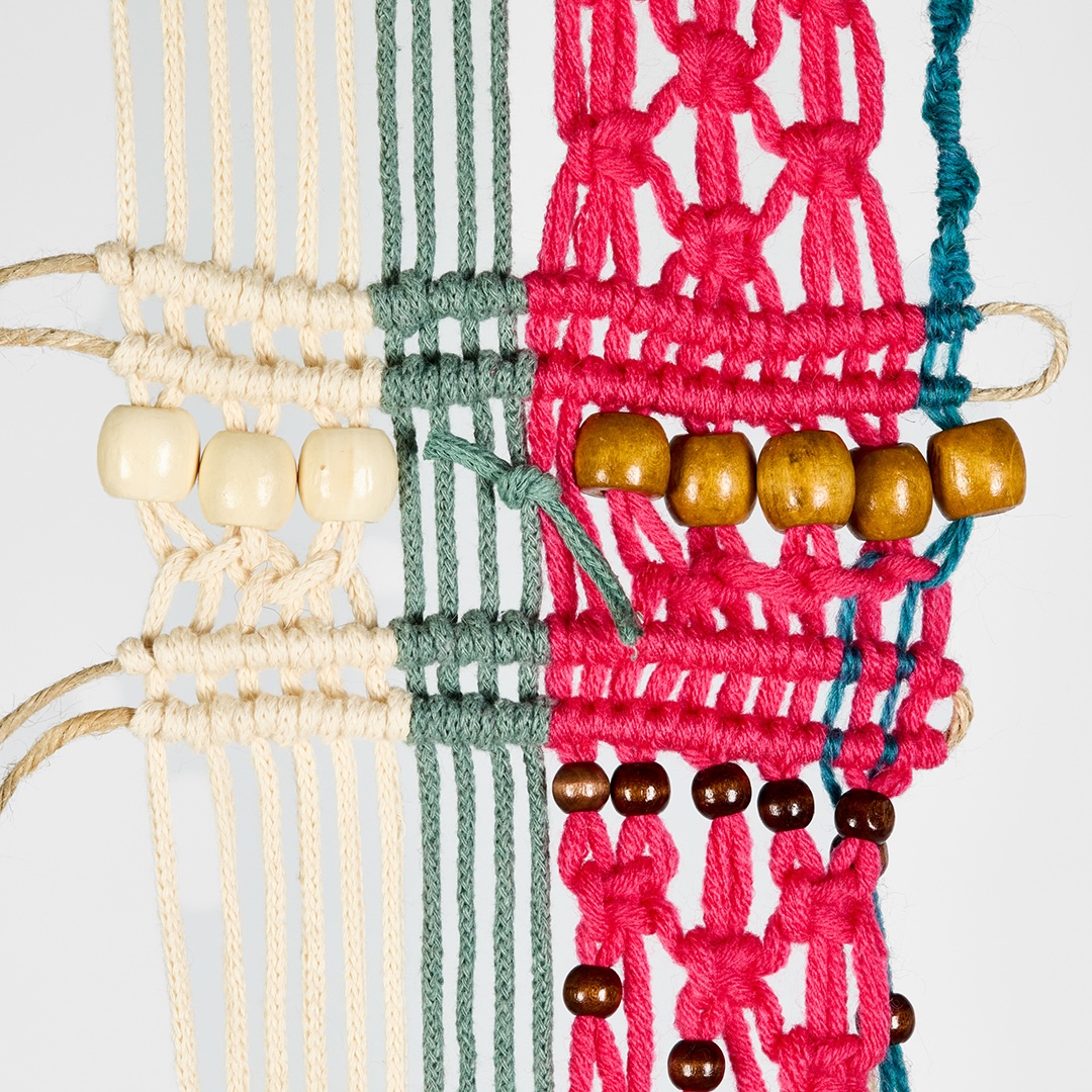 A colourful macramé wall hanging with cream, green, pink and teal cords, decorated with wooden beads against a plain white background.