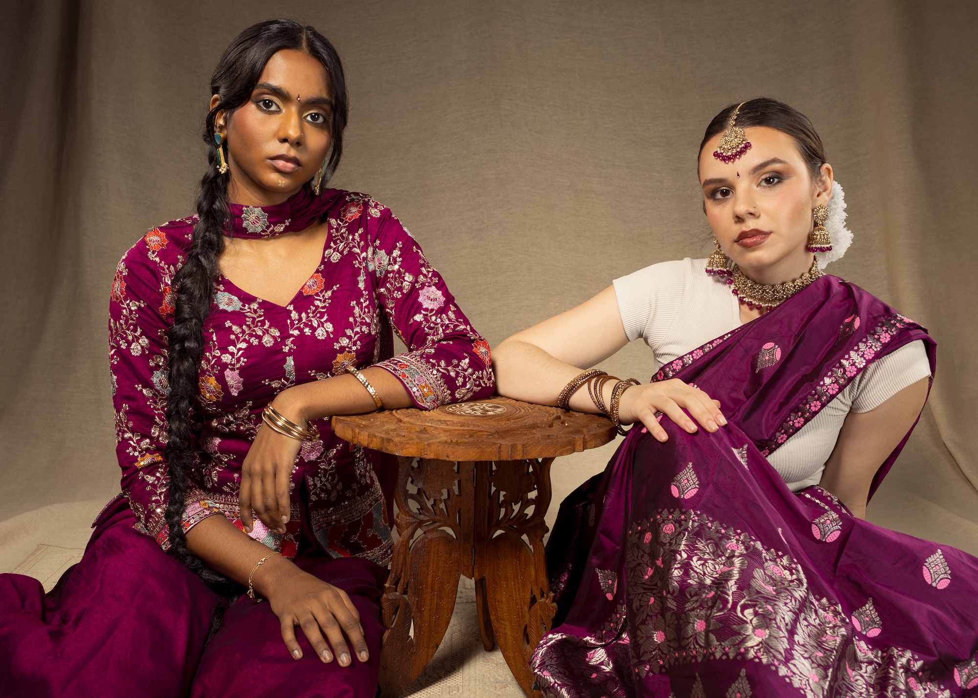 Two fashion models wearing bold purple and white Indian-style garments, pose for a photo while sitting either side of an ornate wooden table.
