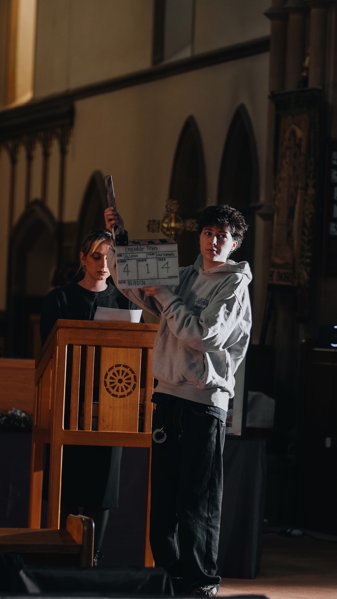 A young man in a grey hoodie holds a clapperboard while standing next to a woman in a black top. They are in a church setting, with wooden architecture and stained glass windows visible in the background.
