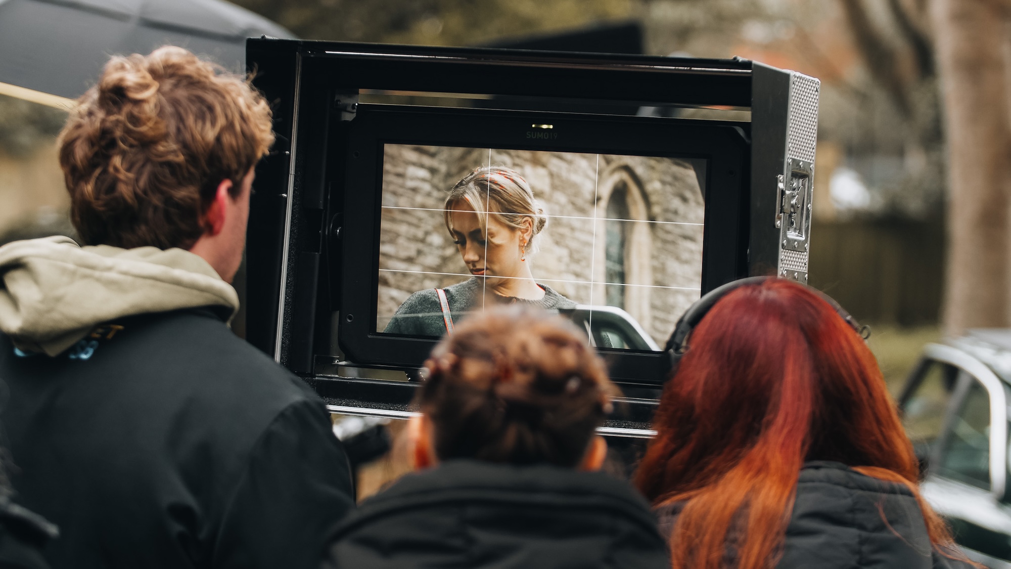 A group of people, including a person with curly hair, watch a monitor displaying a scene featuring a woman with light brown hair in front of a stone building. The setting appears to be an outdoor film shoot with trees in the background.