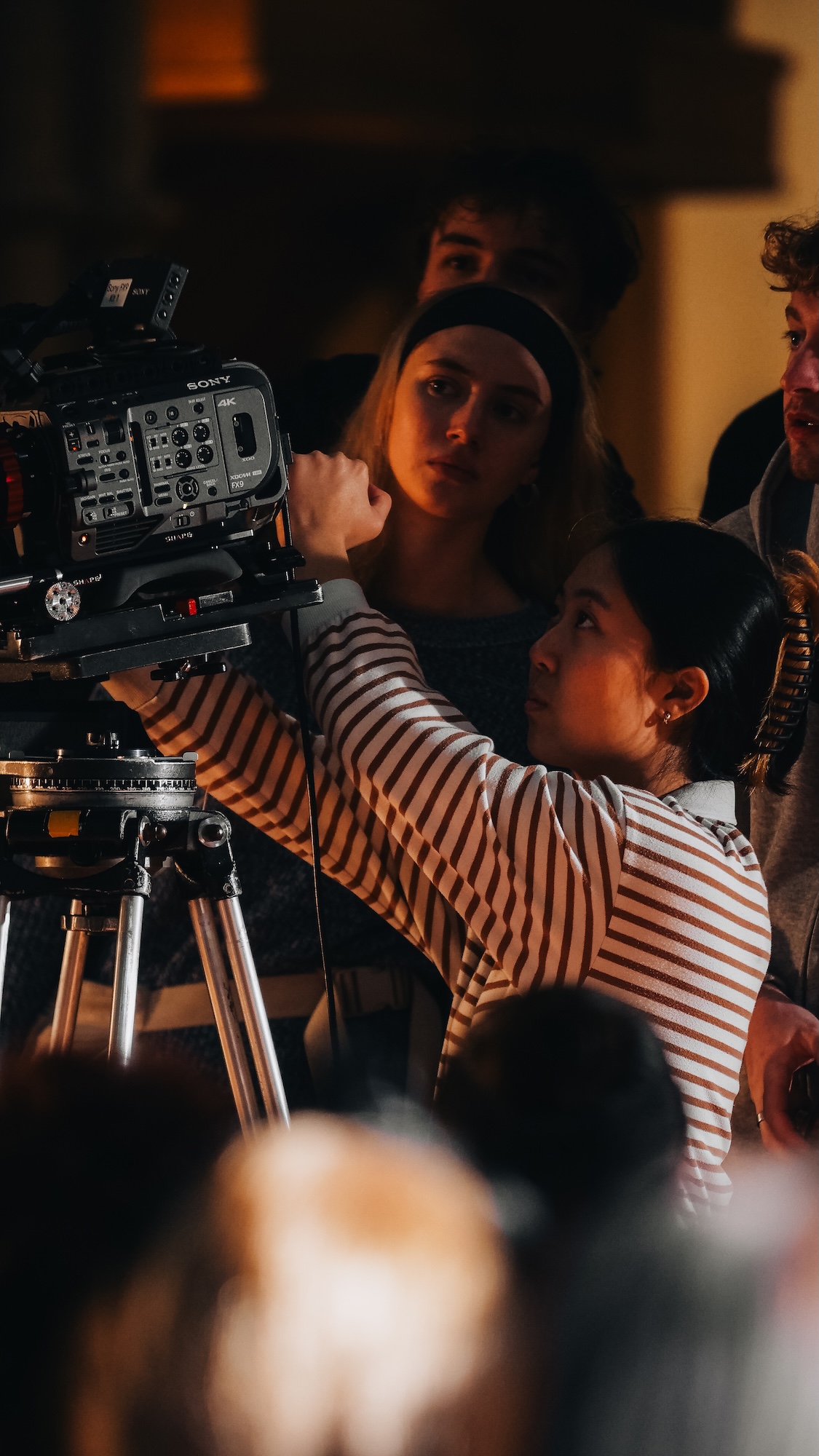 A woman adjusting a film camera on a tripod, wearing a striped shirt. Behind her, a group of people, including another woman in a headband, observe the setup, set against a softly lit background.