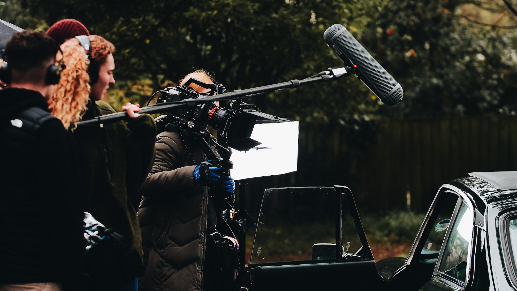 A film crew is set up outdoors, with a woman holding a camera on a tripod and a sound operator using a boom mic. Another crew member is adjusting equipment near a vintage car, while trees create a blurred background.