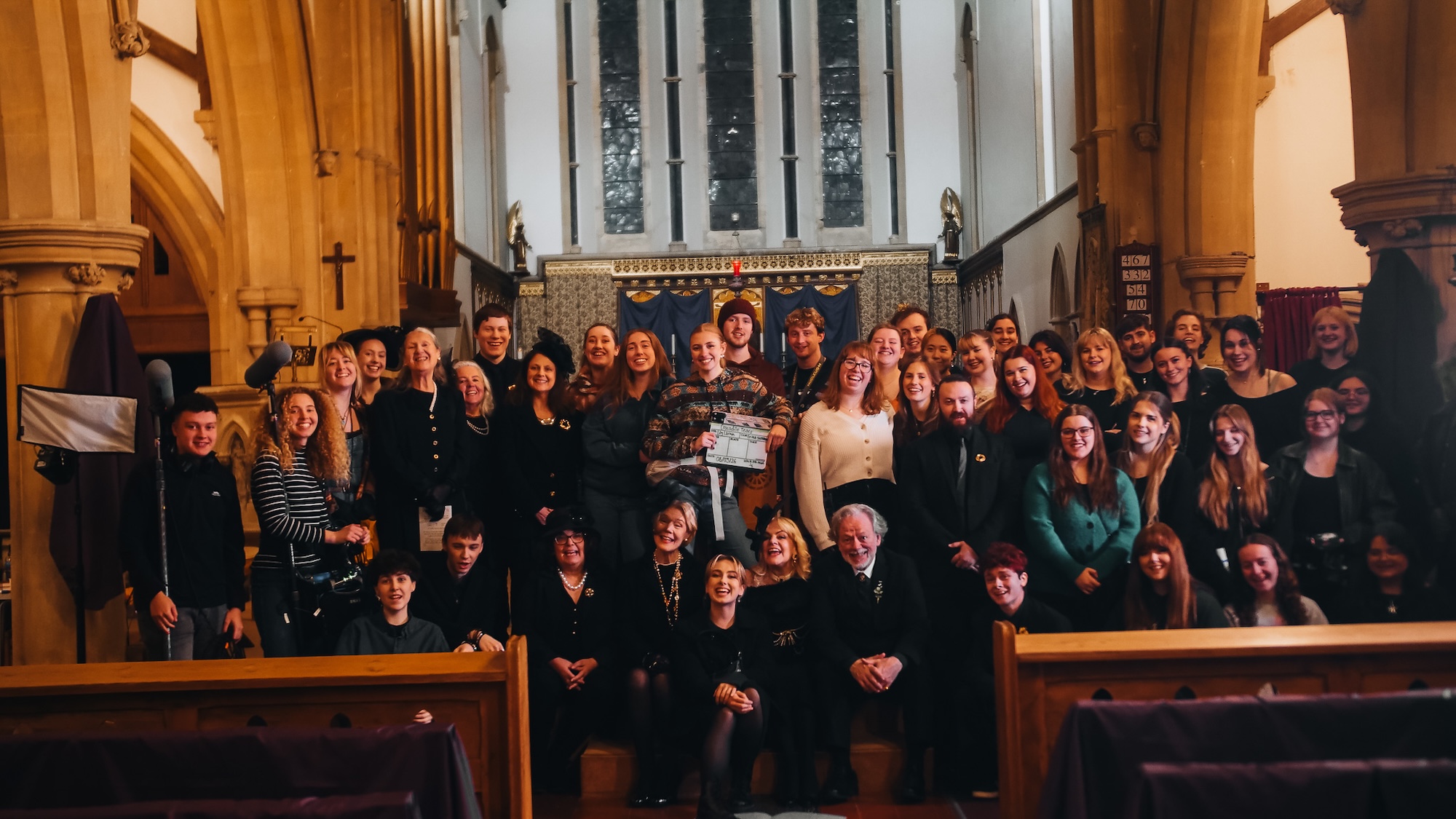 A large group of around 60 people posing together in a church interior. The setting features wooden beams, stained glass windows, and a decorated altar in the background. Individuals are dressed in formal and casual attire, standing and sitting on wooden benches.