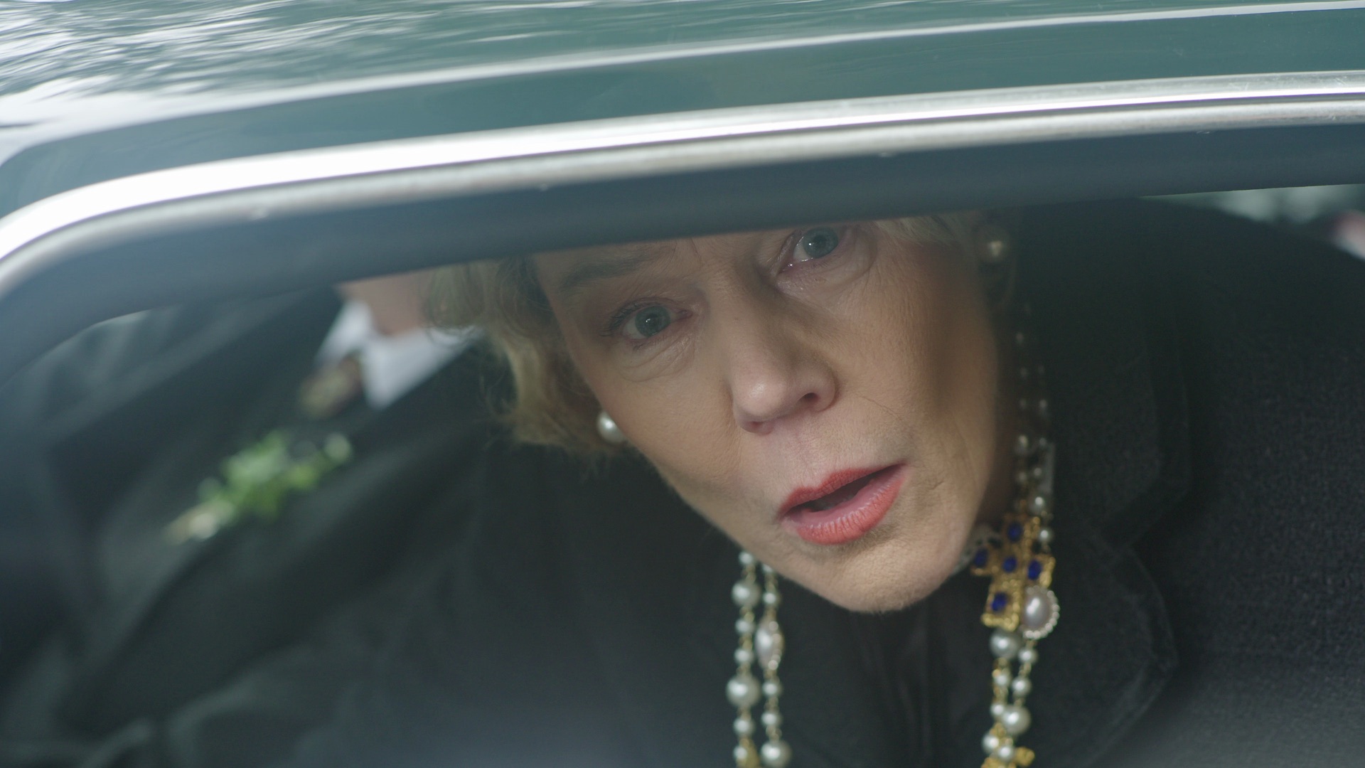 A woman with short blonde hair and pearl necklace leans out of a car window, looking forward. She is wearing a black coat, and the background includes blurred grey figures and greenery.