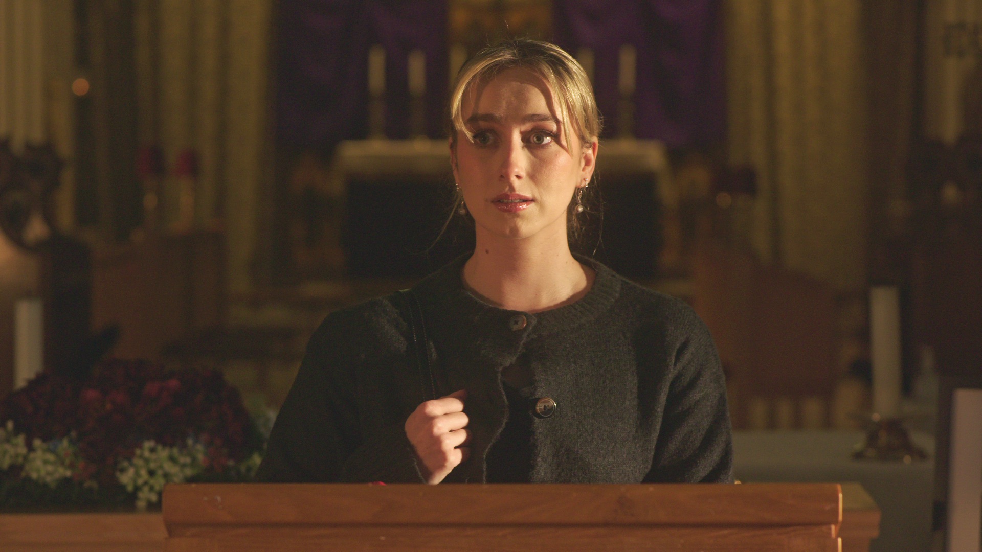 A young woman with light brown hair, styled in a ponytail, stands at a wooden lectern in a church. She wears a dark cardigan and is positioned in front of a softly illuminated altar, with purple drapes and floral arrangements in the background.