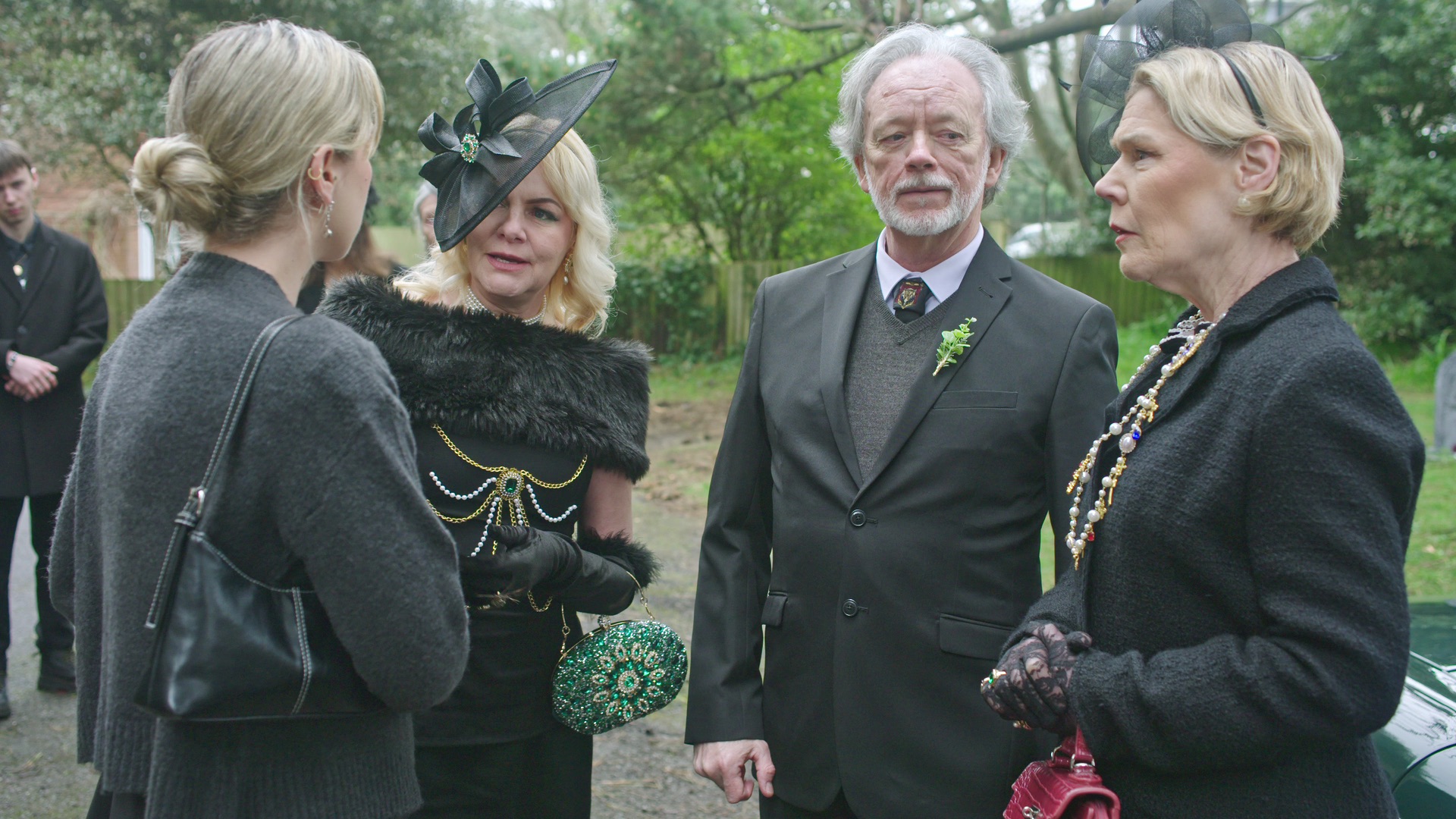 Four adults gather outdoors, engaged in conversation. One woman wears a black dress adorned with a feathered hat, while another in a dark coat and silver necklace looks on. A man in a suit with a small green boutonniere stands between them, alongside a woman with a black handbag.