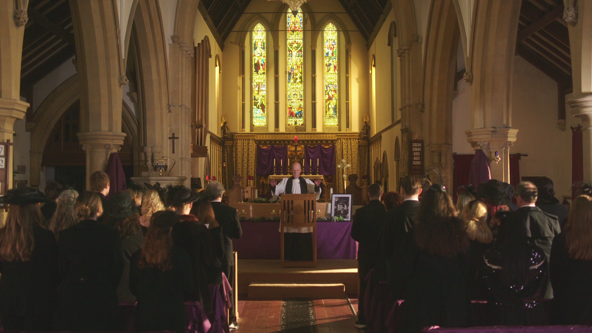 A church interior during a funeral service. Attendees in black clothing are seated, facing a person at the front by an altar, which is adorned with purple cloth. Stained glass windows illuminate the setting with colourful light.