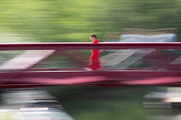 Motion-blurred image of person in red dress on burgundy boat moving across water with green shoreline background.