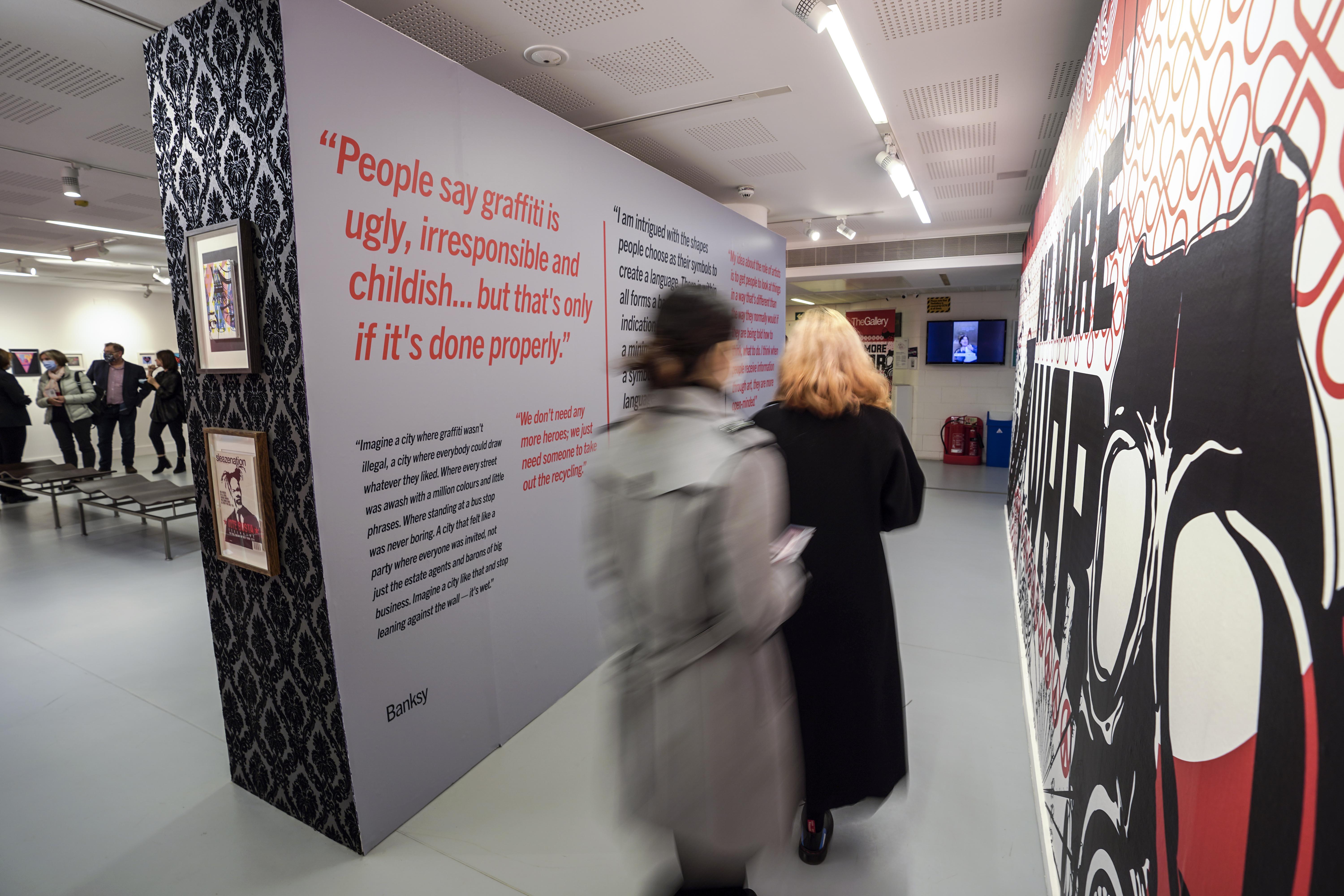 Two people walking down a gallery walkway. There is a large artwork printed on the right-hand wall, and blocks of text printed on the left-hand side.
