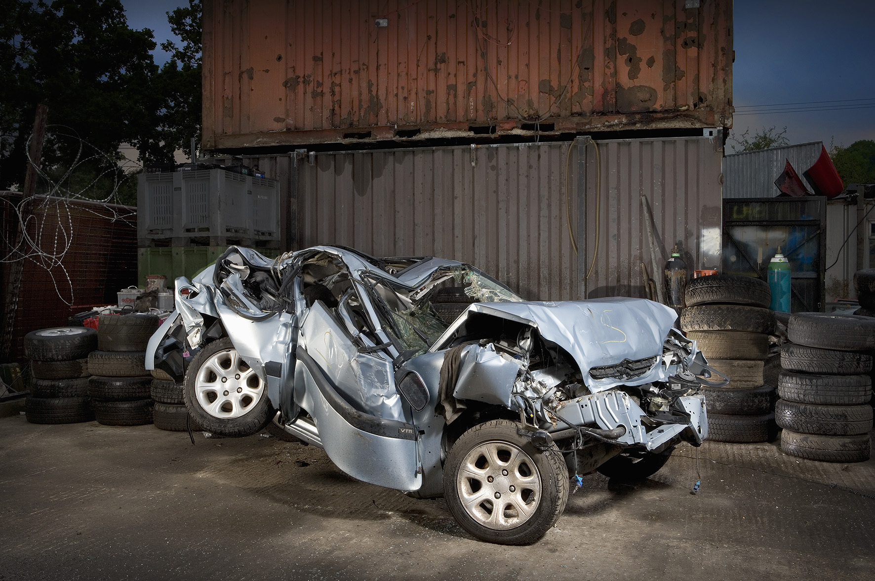 A crumpled car lying with metal shipping containers and stacks of tyres in the background.