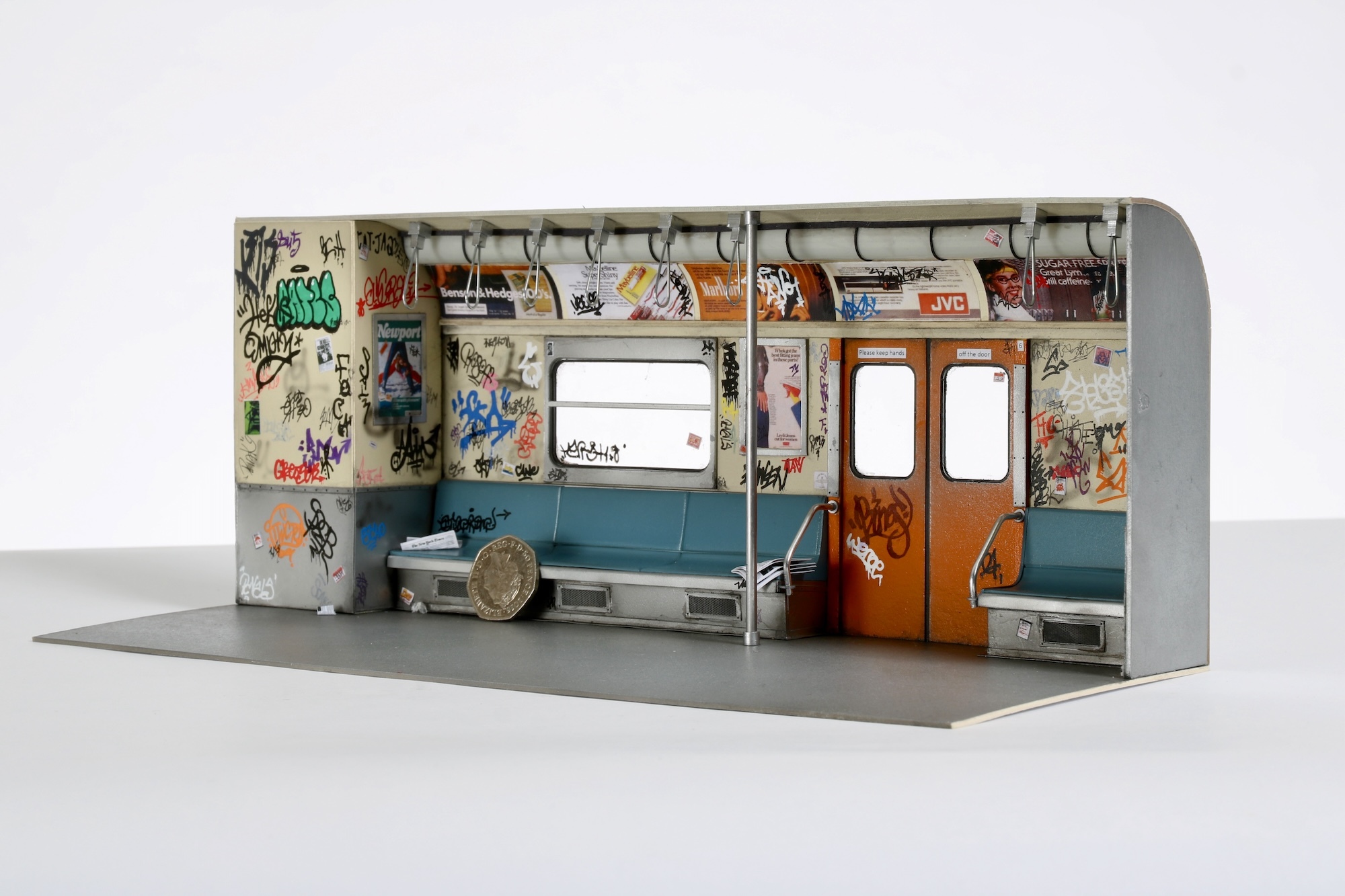 Model of the interior of a London tube carriage with seats, graffitied walls and overhead adverts.