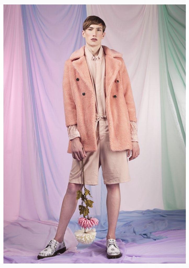 Image of a man in a pink coat and shorts holding a pair of flowers, which are pointing downwards.